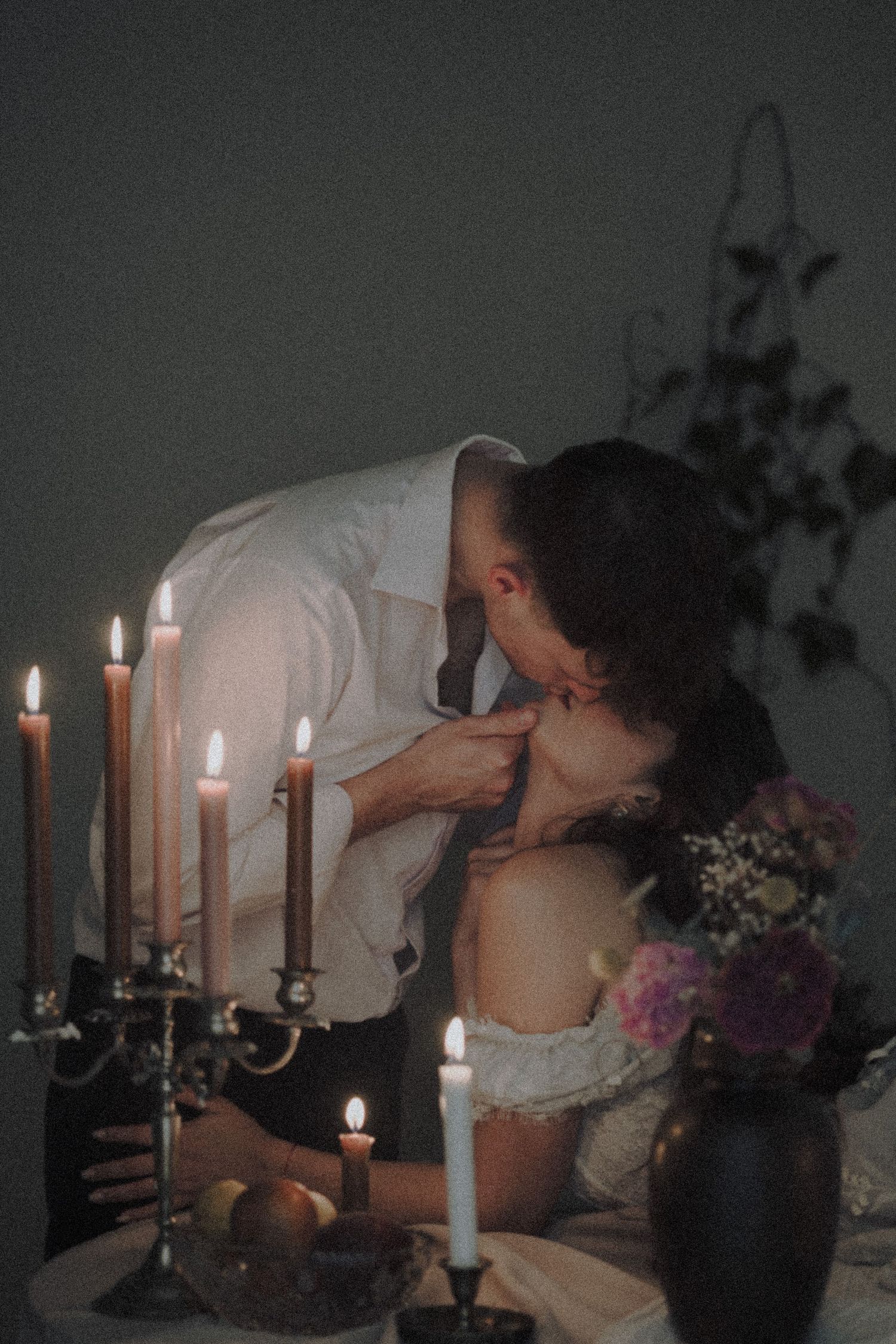 Romantic candlelit scene with a couple embracing, surrounded by glowing candles and flowers in a moody, dimly lit setting.