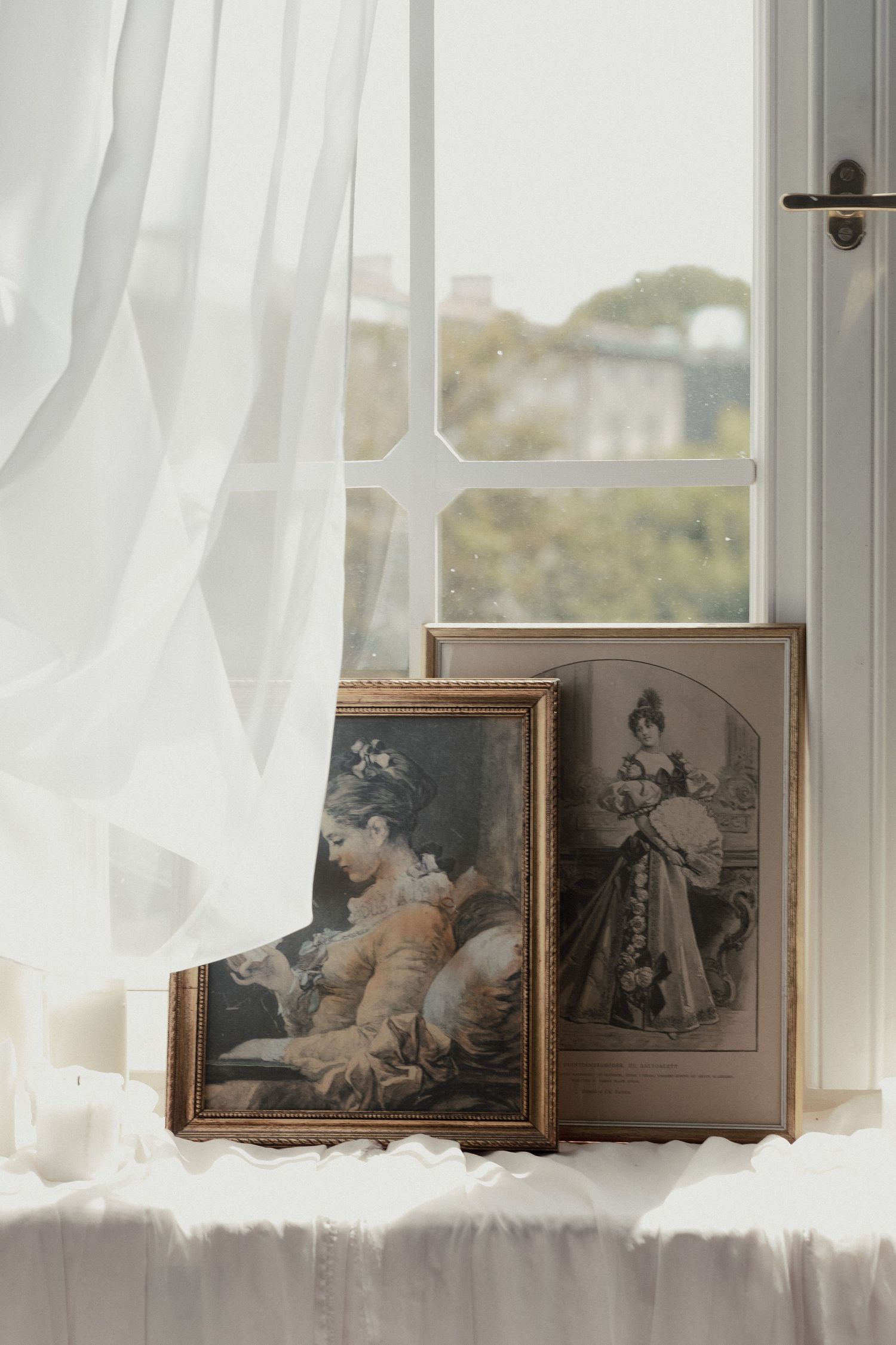 Vintage framed artwork and photographs rest on a windowsill with sheer white curtains and natural light streaming in.