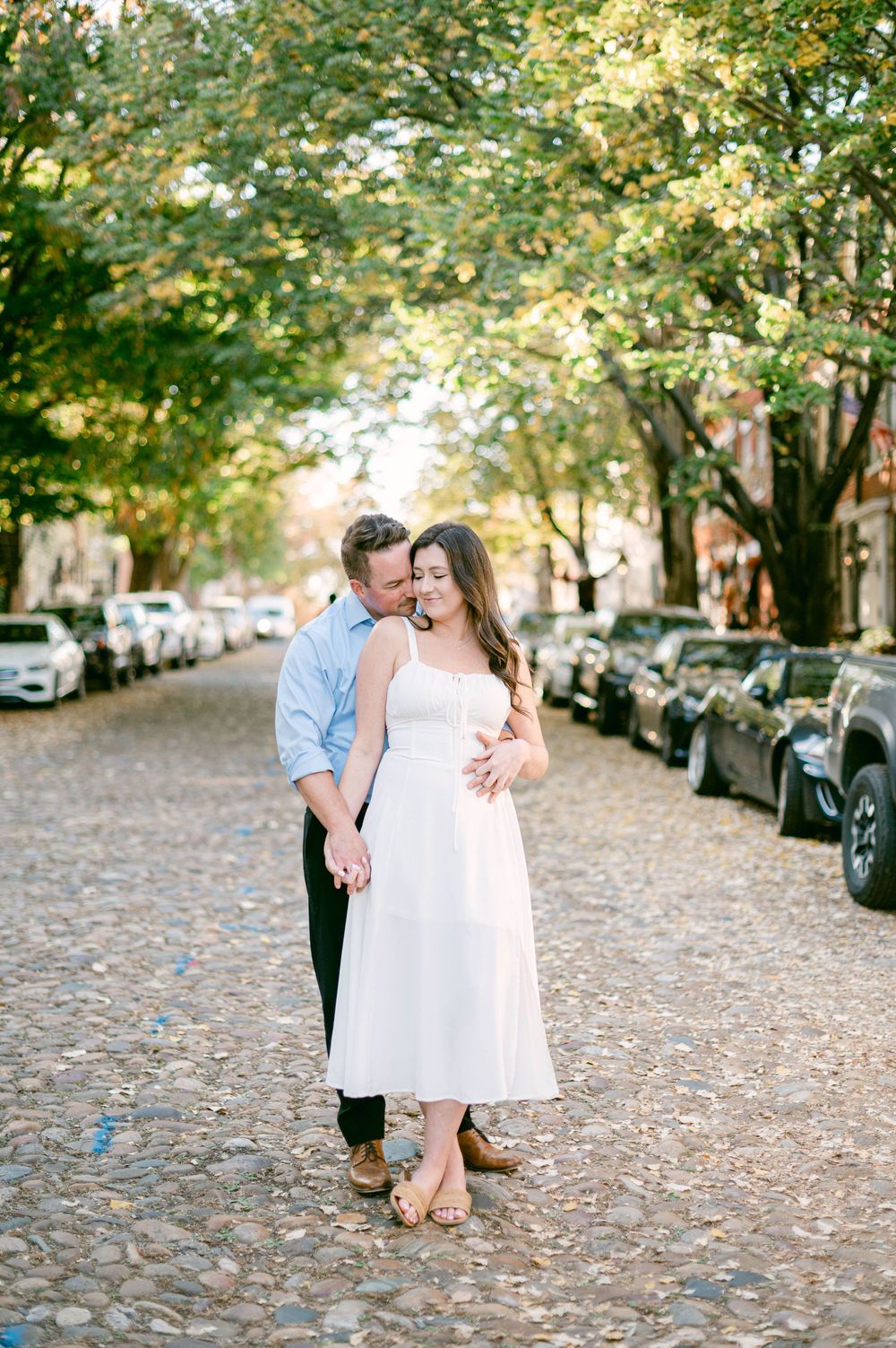 Couple embracing on tree-lined cobblestone street with parked cars.