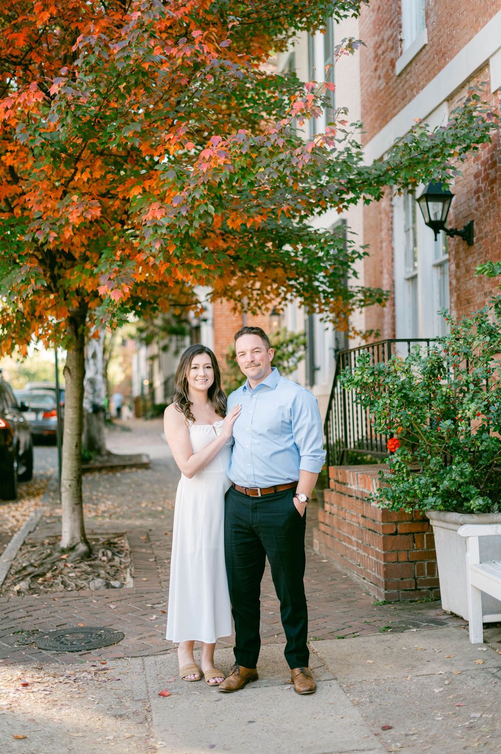 Couple poses together on a brick sidewalk with autumn foliage and historic architecture.