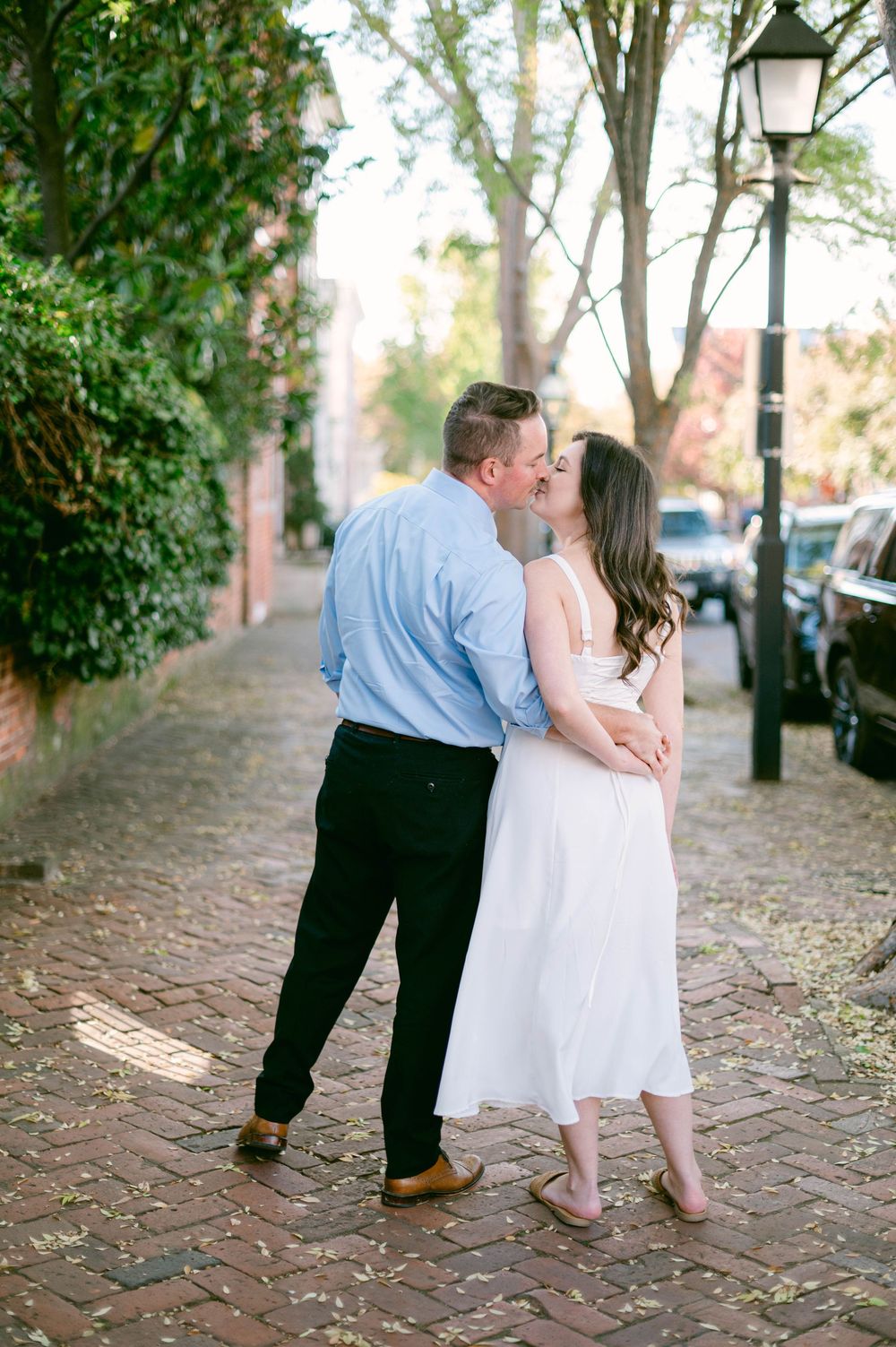 Couple sharing a romantic kiss on a cobblestone path near vintage lampposts.