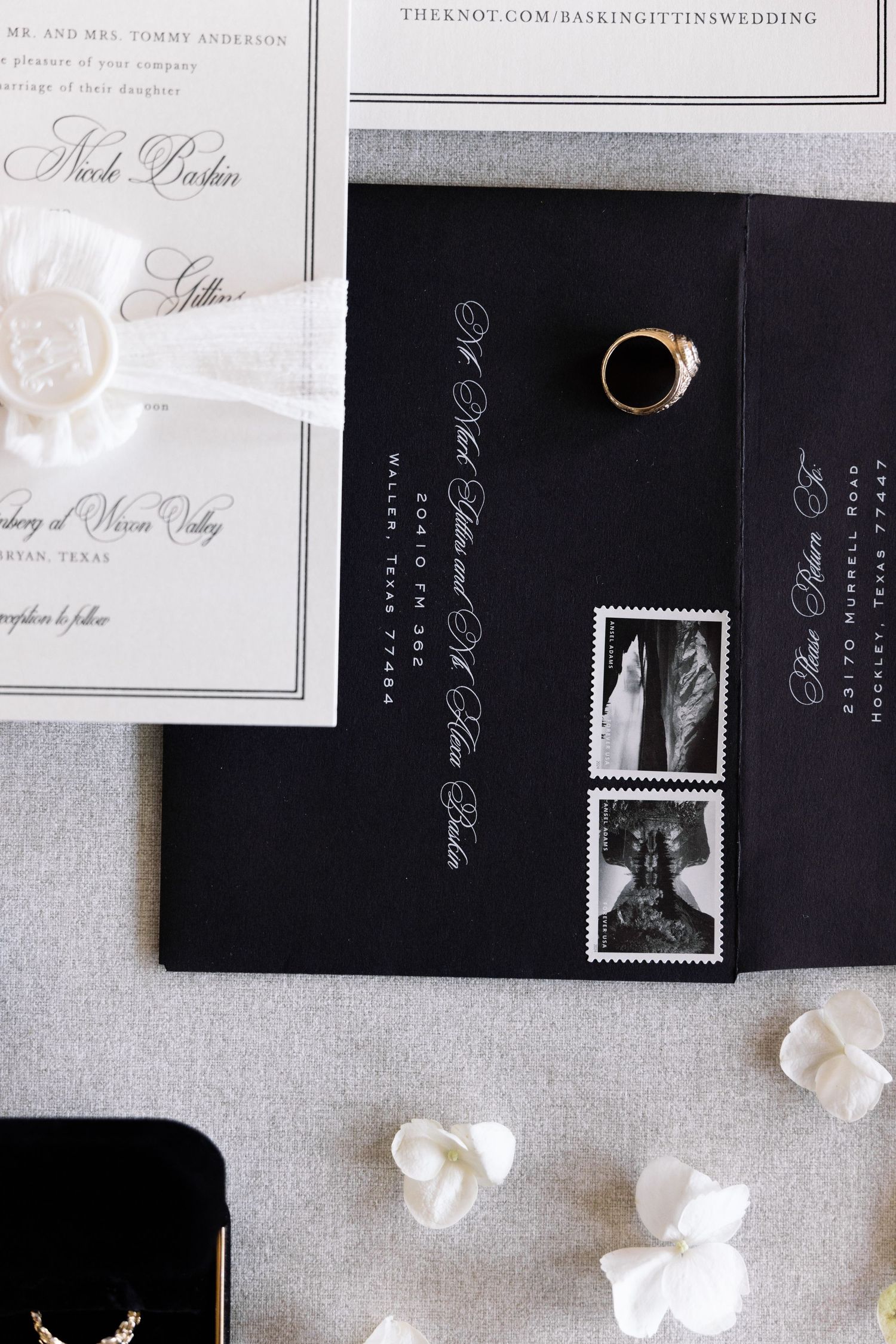Elegant black and white wedding invitation suite with delicate paper flowers scattered on a gray textured background.