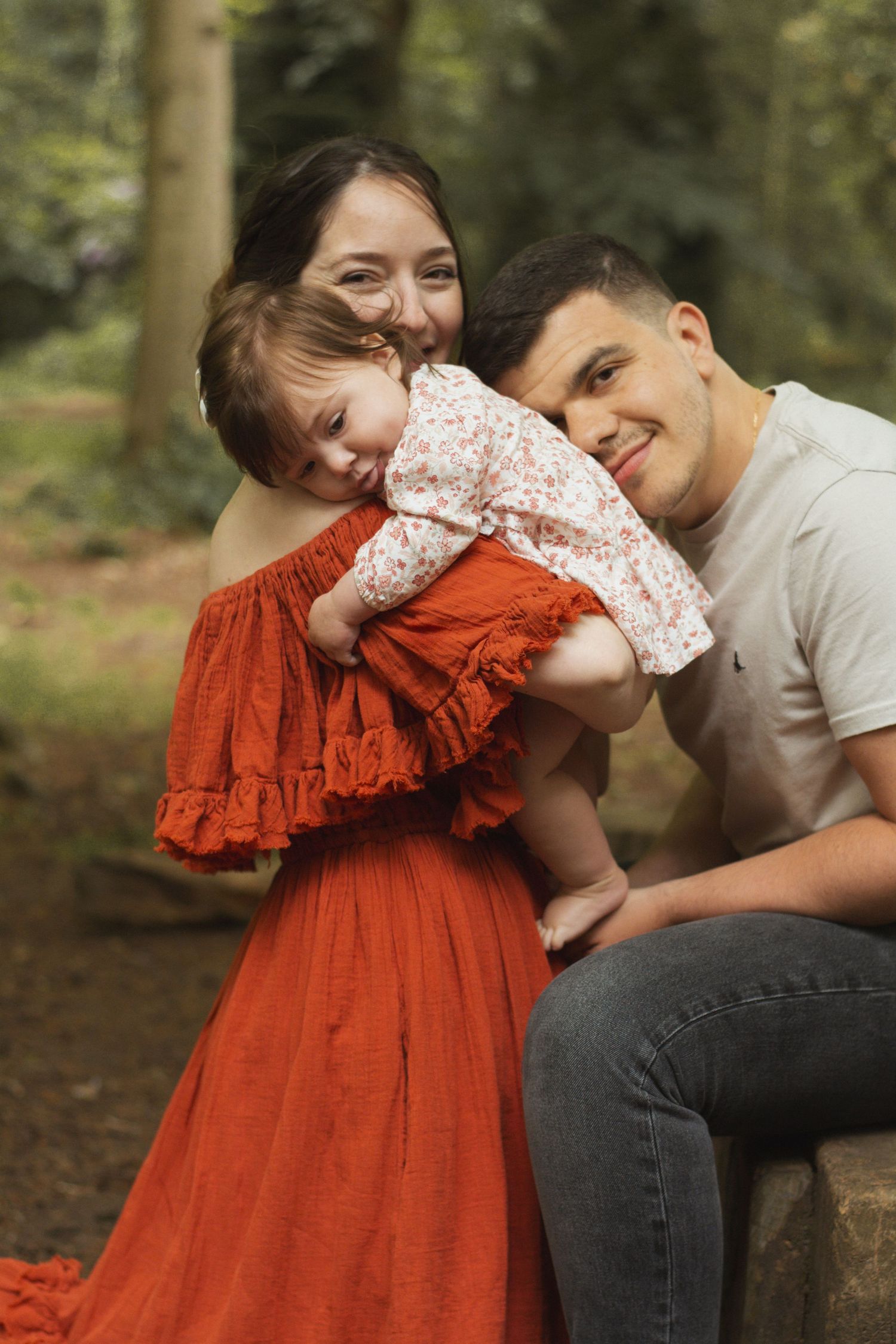 A family shares a joyful moment together in a forest setting, with the mother wearing a flowing red dress.
