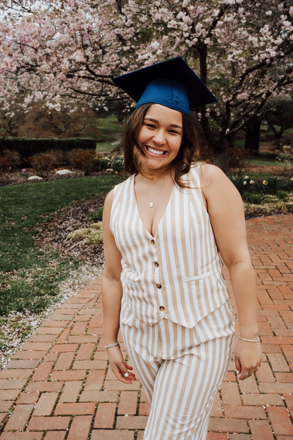 A graduate wearing a blue cap and striped outfit poses near blooming cherry blossom trees on a brick pathway at Hershey Gardens.