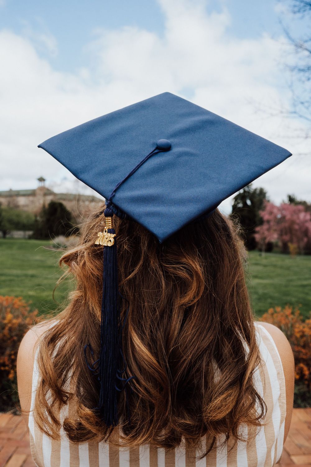 A blue graduation cap worn by high school graduate with long wavy brown hair standing outdoors on a spring day at Hershey Gardens.
