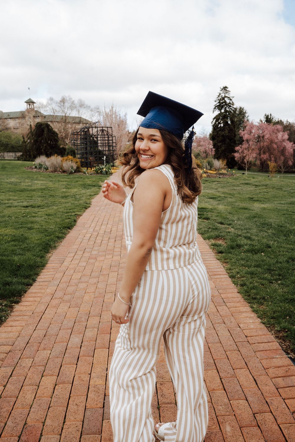 A high school graduate wearing her cap poses on a brick pathway with campus buildings and trees in the background at Hershey Gardens.