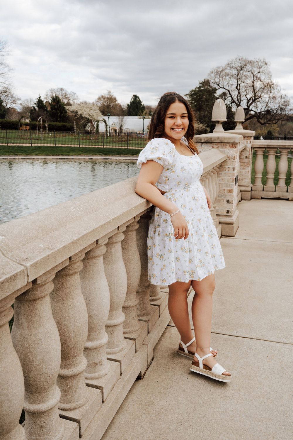 High school senior at lifestyle photoshoot wearing a white floral dress posing along a stone balustrade by a lake at Hershey Gardens.