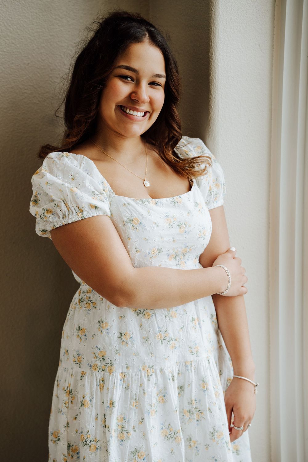 High school senior wearing a floral white dress with puff sleeves for senior portrait session by a window at Hershey Gardens.