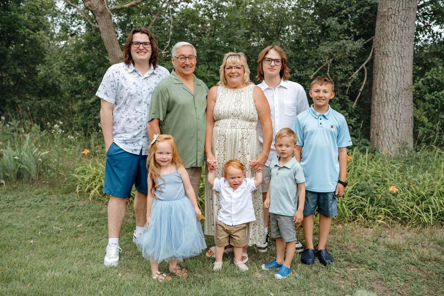 A large family group poses together outdoors on a sunny day with trees and greenery in the background.