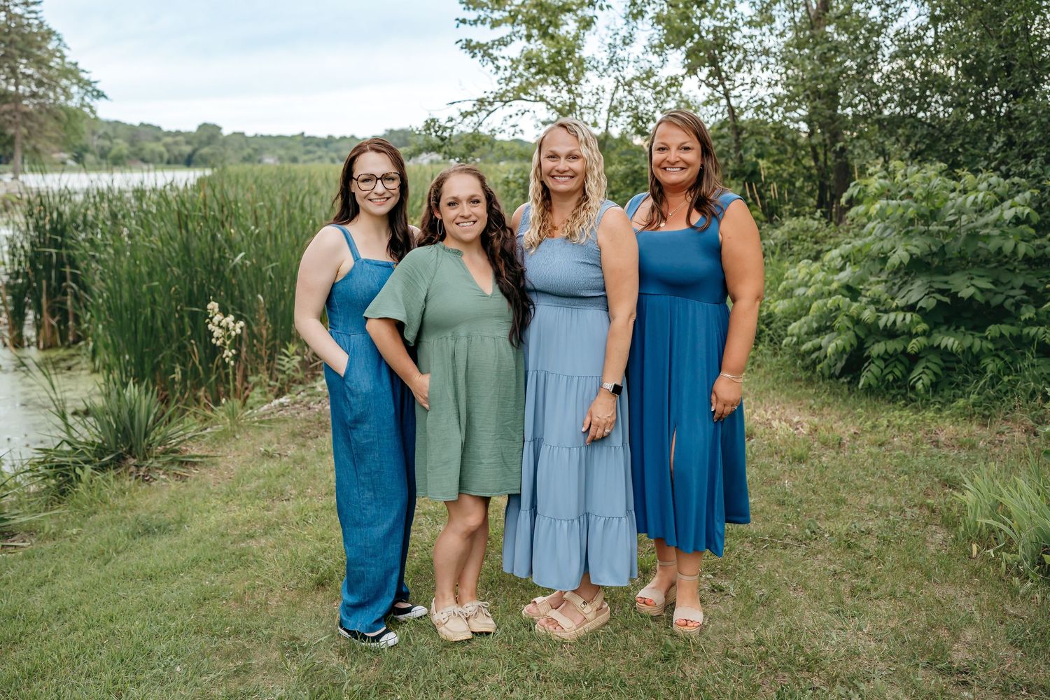 Four friends wearing coordinating blue and green dresses stand together by a lake on a summer day.
