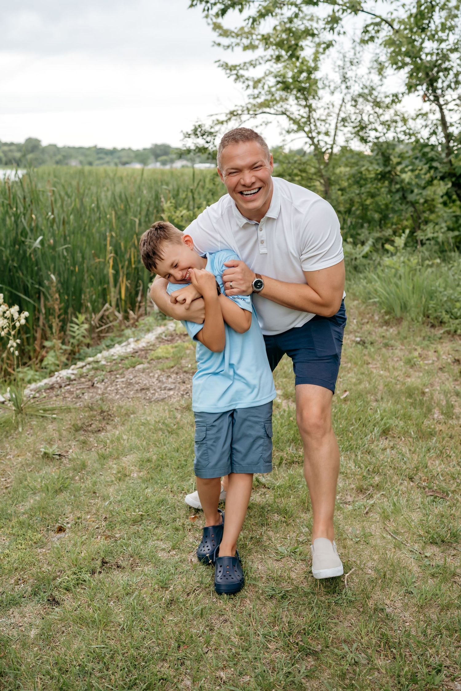 Parent and child playing and laughing together on a grassy path near water.