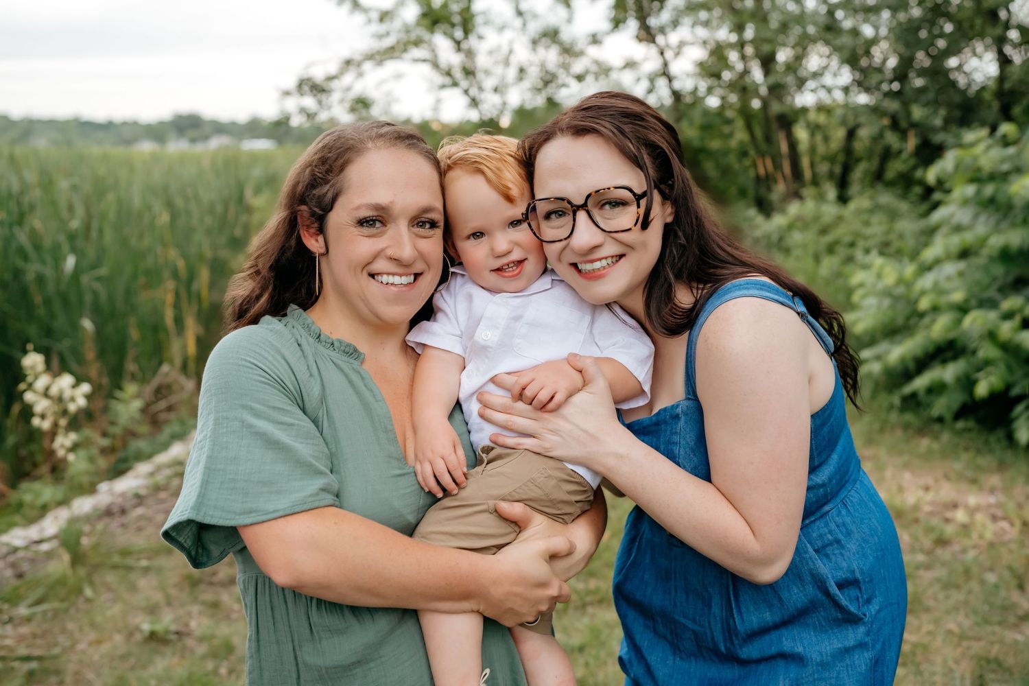Three people share a tender moment together outdoors against a natural backdrop of greenery and water.