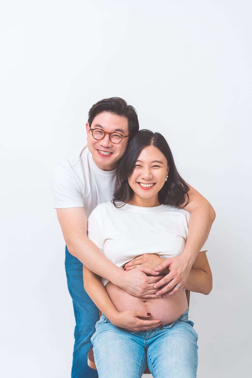 Expectant couple in white shirts embrace against white background as they share a joyful moment during pregnancy.