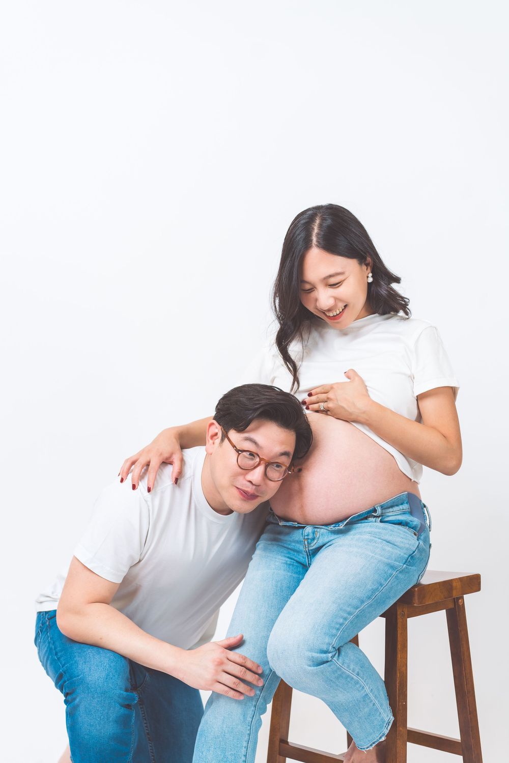 Expecting couple in white shirts and jeans share an intimate moment as they touch a pregnant belly while sitting on a stool.