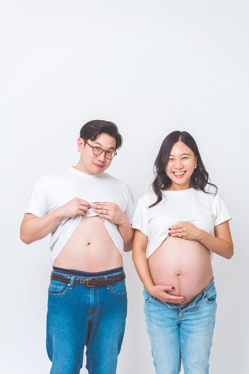 A couple in white t-shirts and jeans playfully mimic pregnancy poses by pushing out their bellies against a white background.