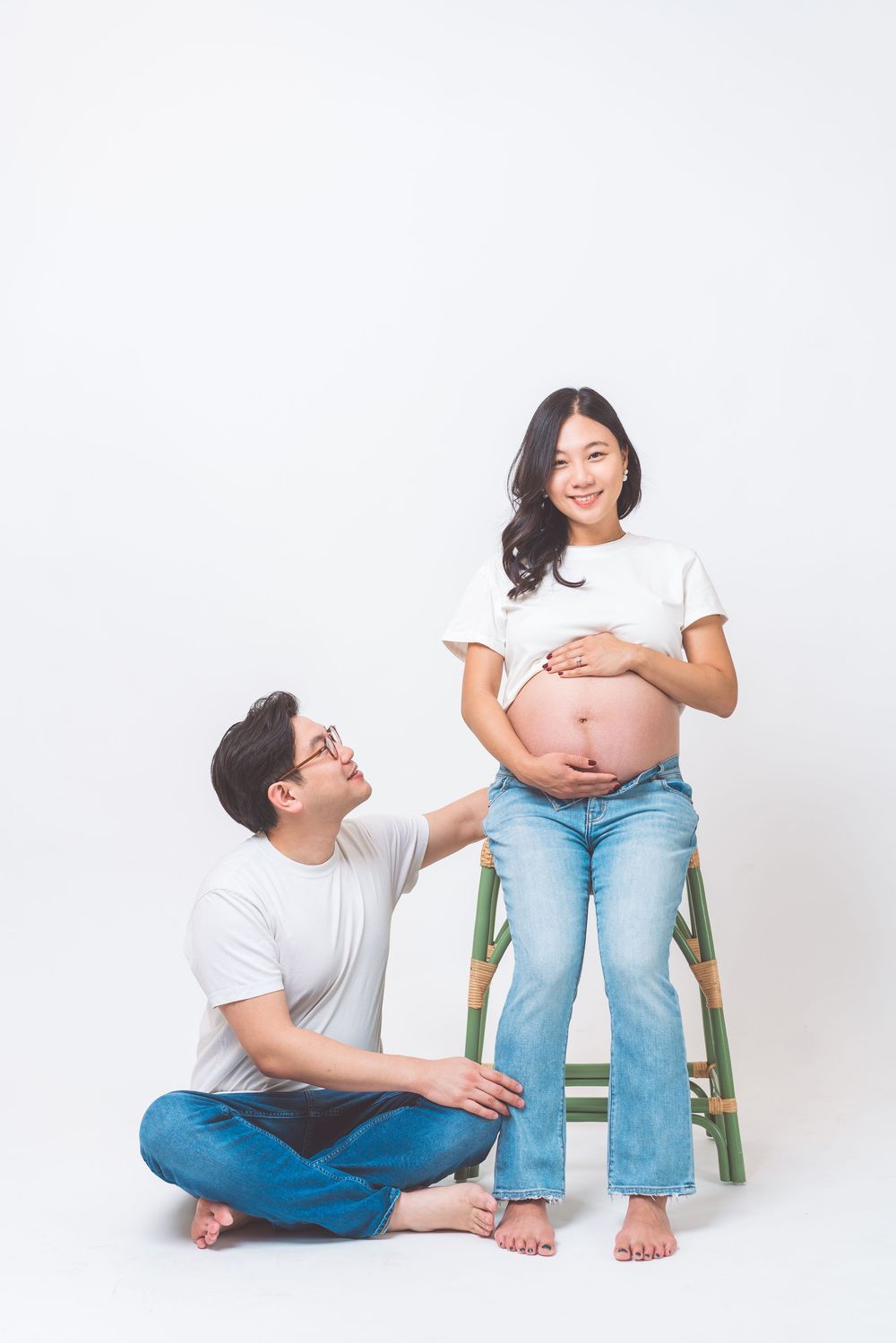 Couple posing for a maternity photo with one seated on floor while the other stands on a wooden stool against white backdrop.