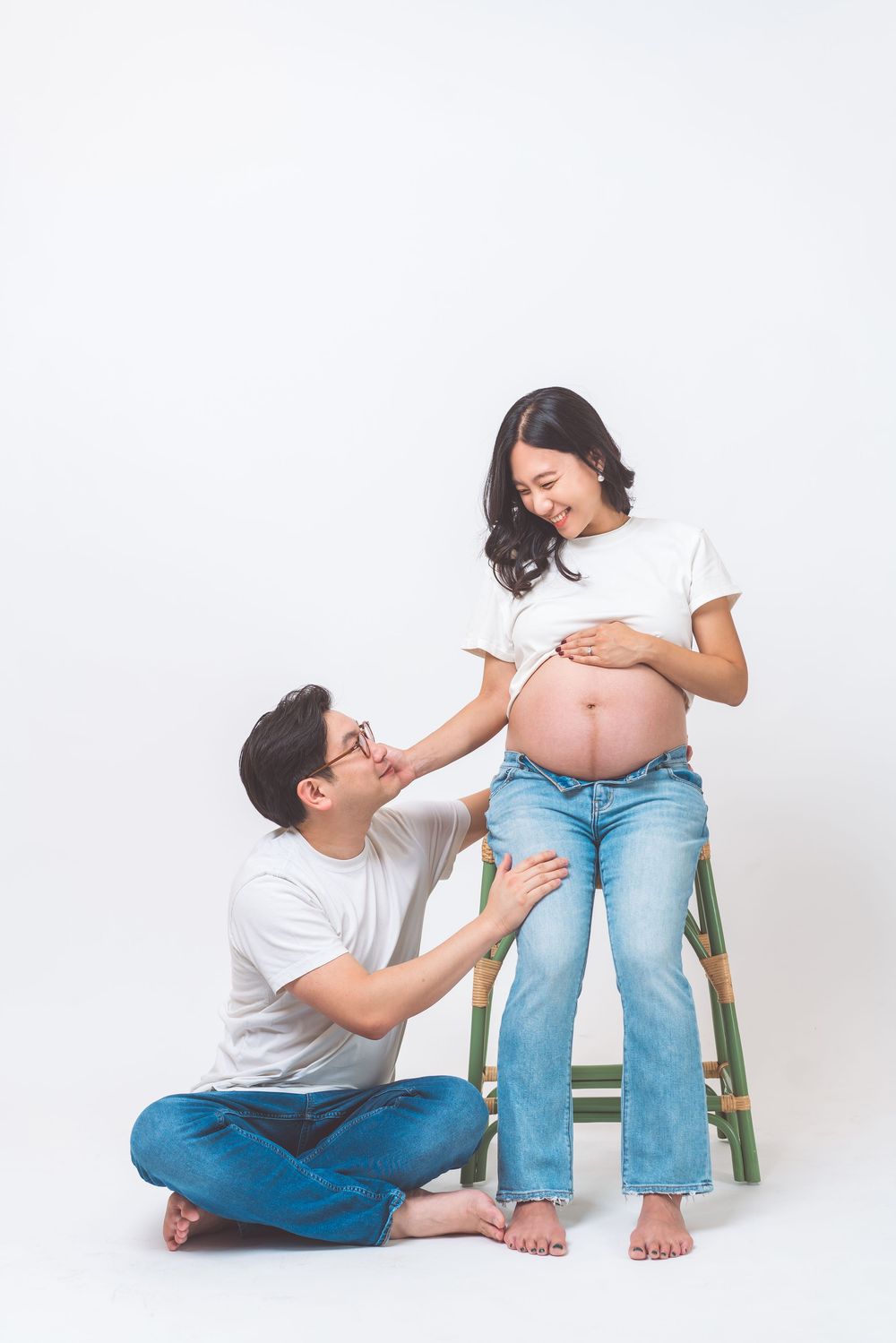 A couple shares a tender moment during maternity photoshoot with the pregnant woman standing by a wooden stool.