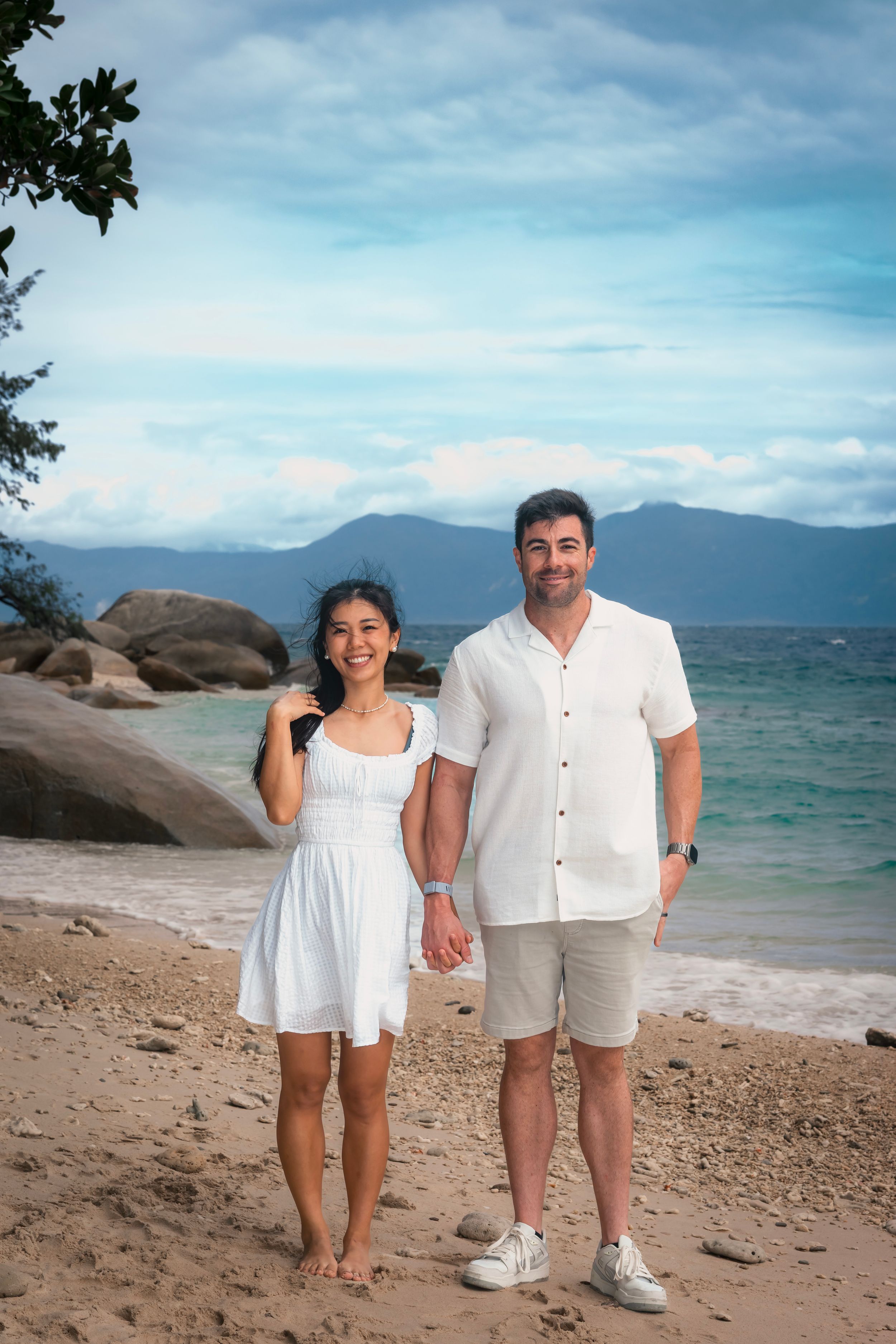 A couple stands together on a tropical beach with mountains and turquoise water in the background.