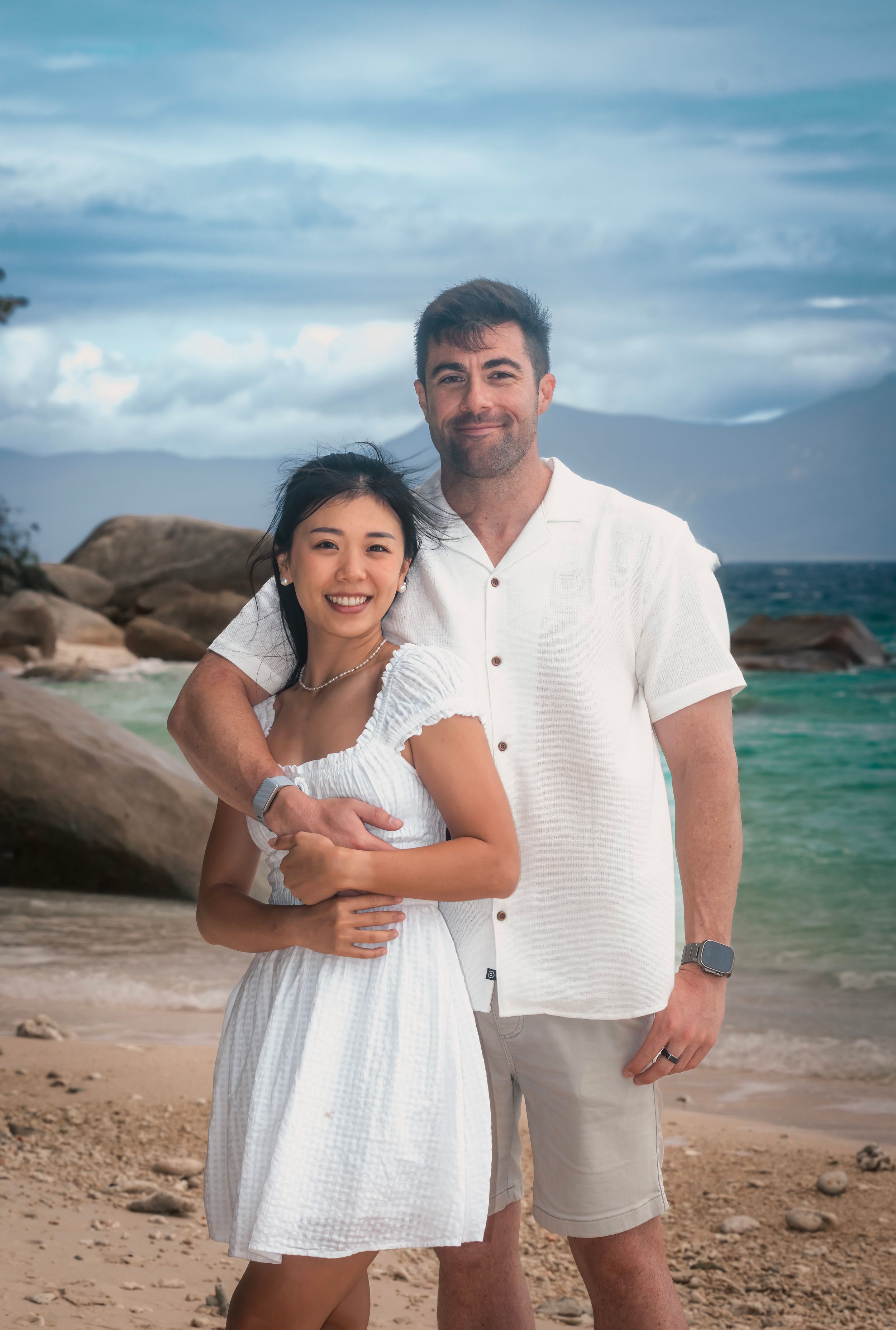 A couple poses together on a scenic tropical beach with turquoise water and rocky coastline in the background.