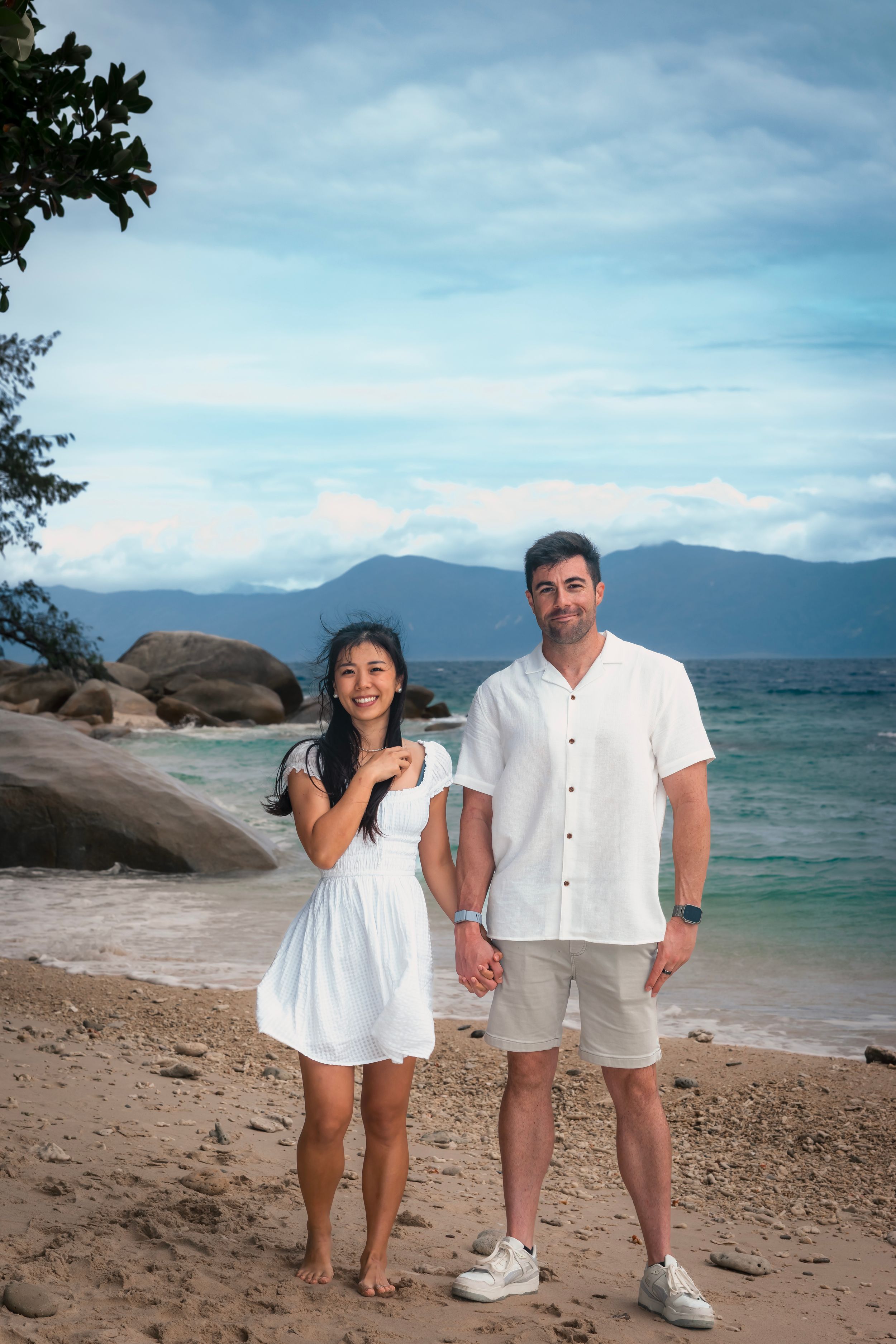 A couple poses together on a tropical beach with rocks and mountains in the background on a sunny day.