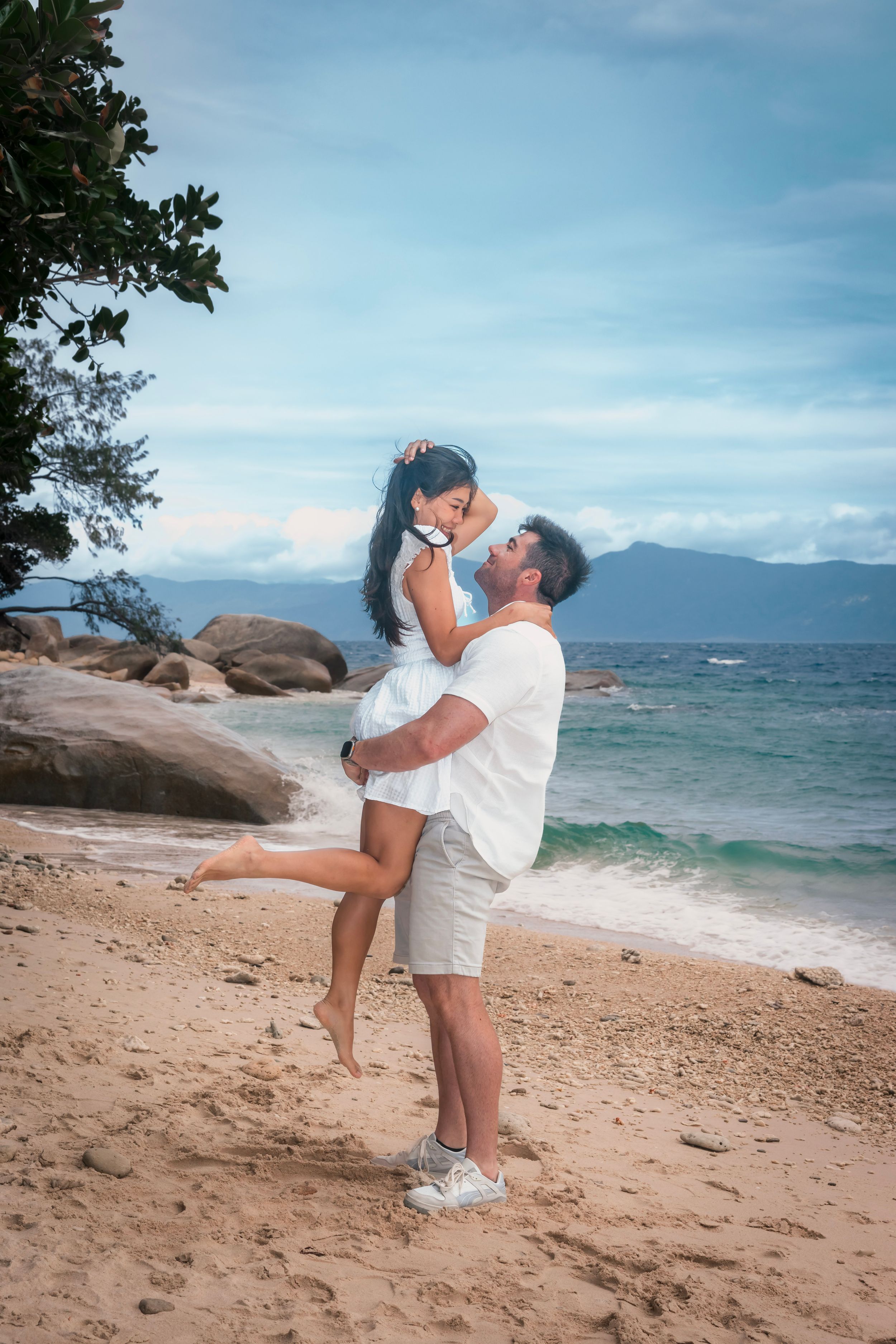 A romantic couple embraces on a tropical beach with rocky coastline and mountains visible in the distance.