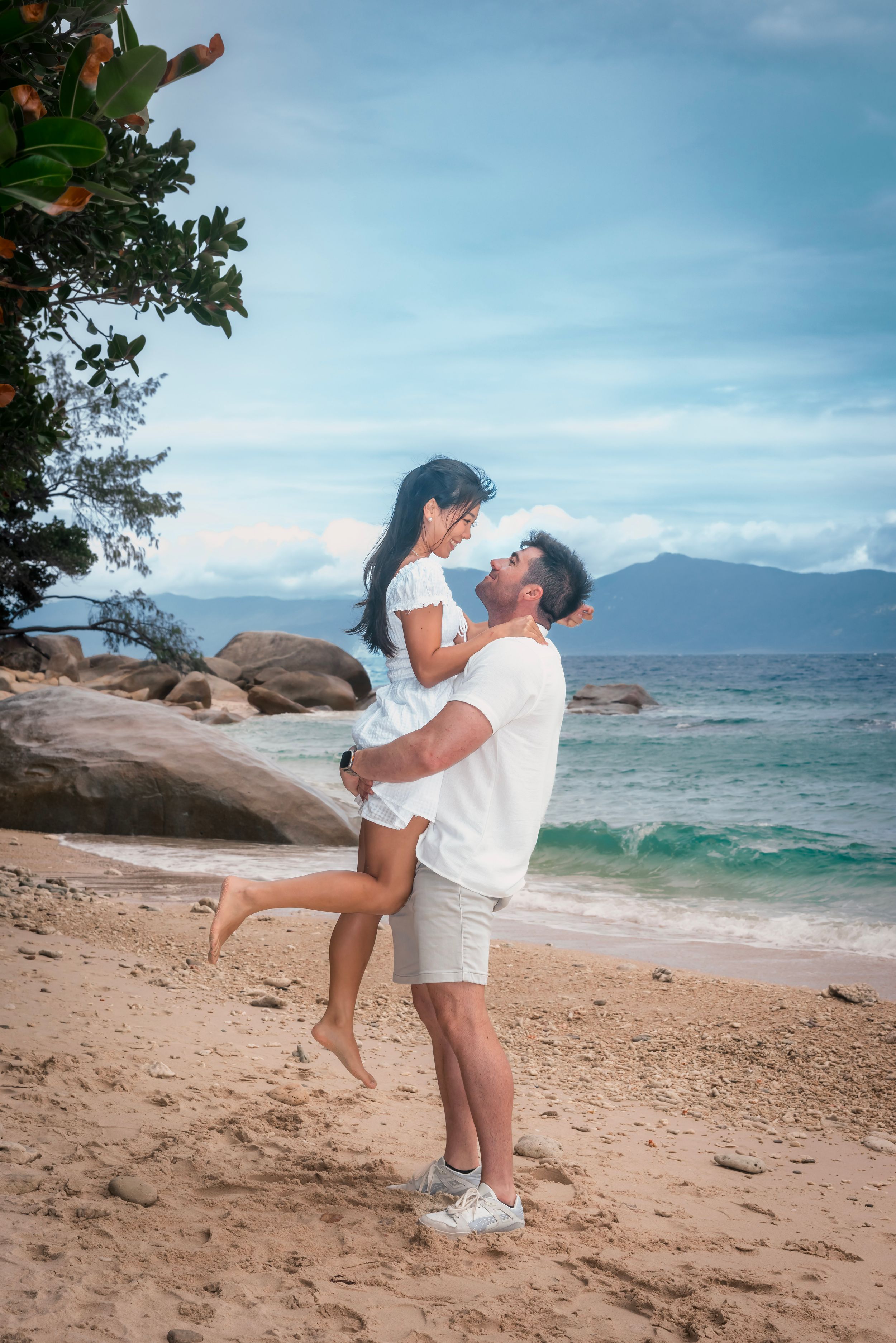 A romantic beach scene with couple embracing on sandy shore with mountains and ocean waves in the background.