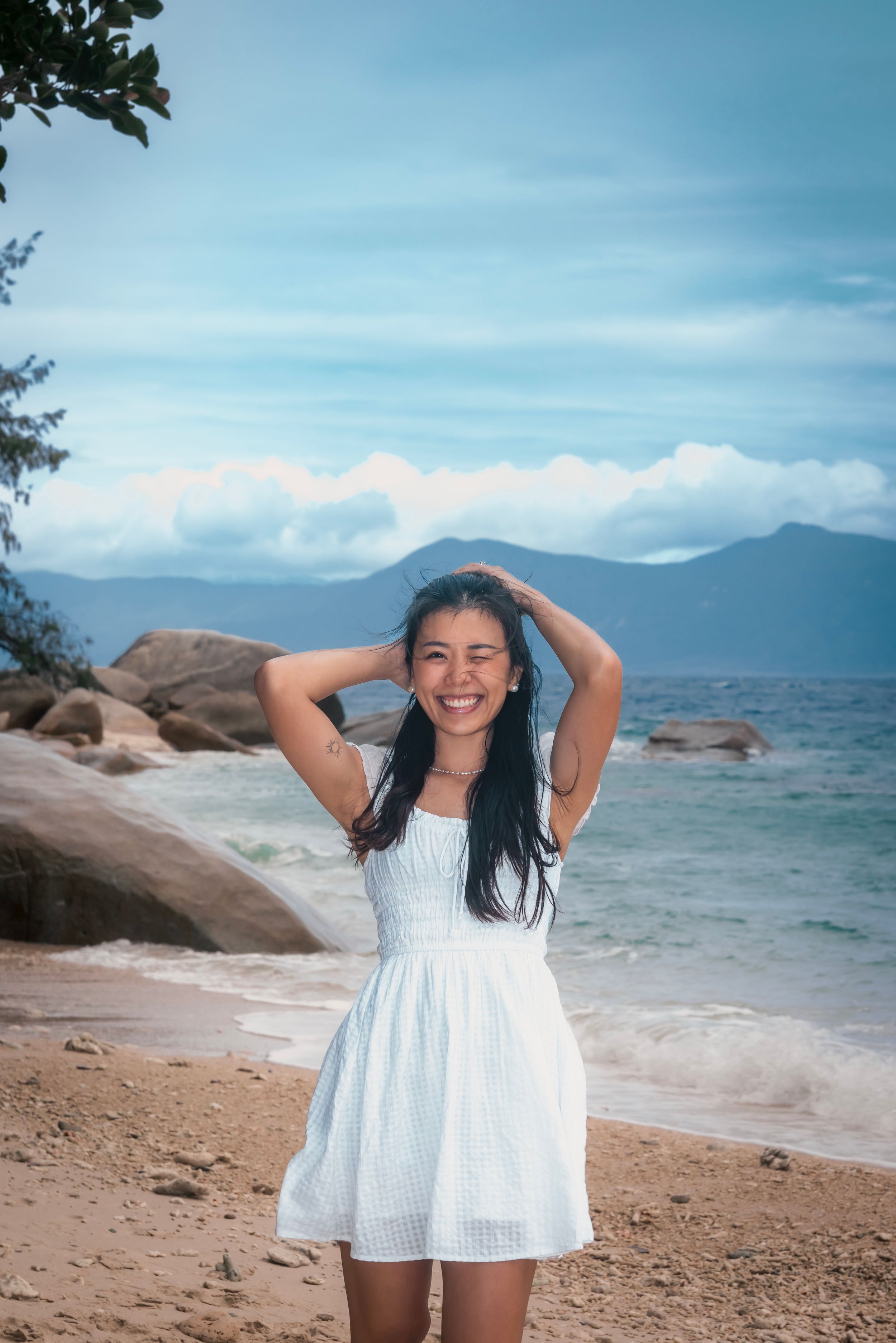 A woman in a flowing white dress poses joyfully on a scenic beach with rocky shores and mountains in the distance.