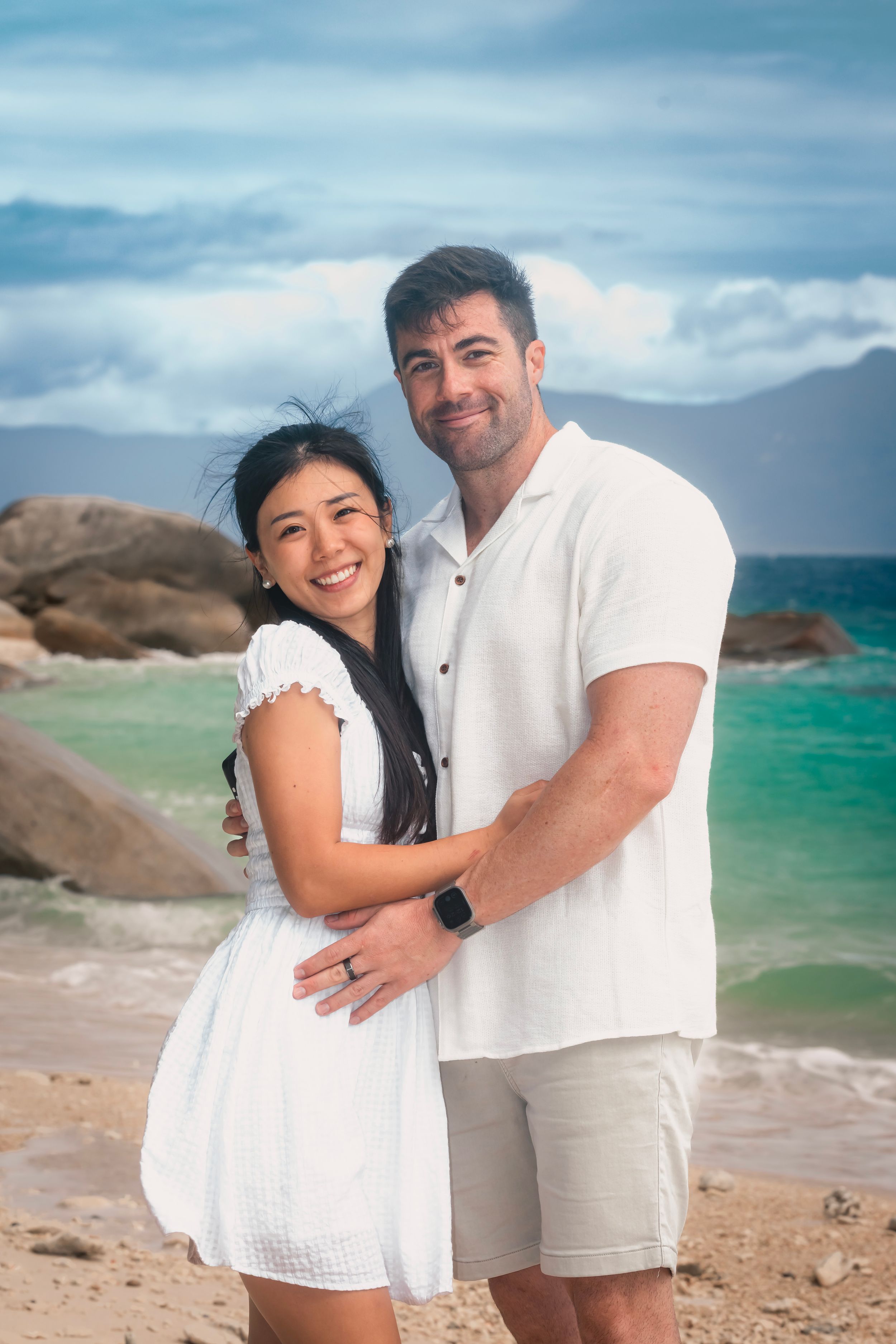 A couple in white outfits shares romantic moments on a rocky beach with turquoise waters and cloudy skies in the background.