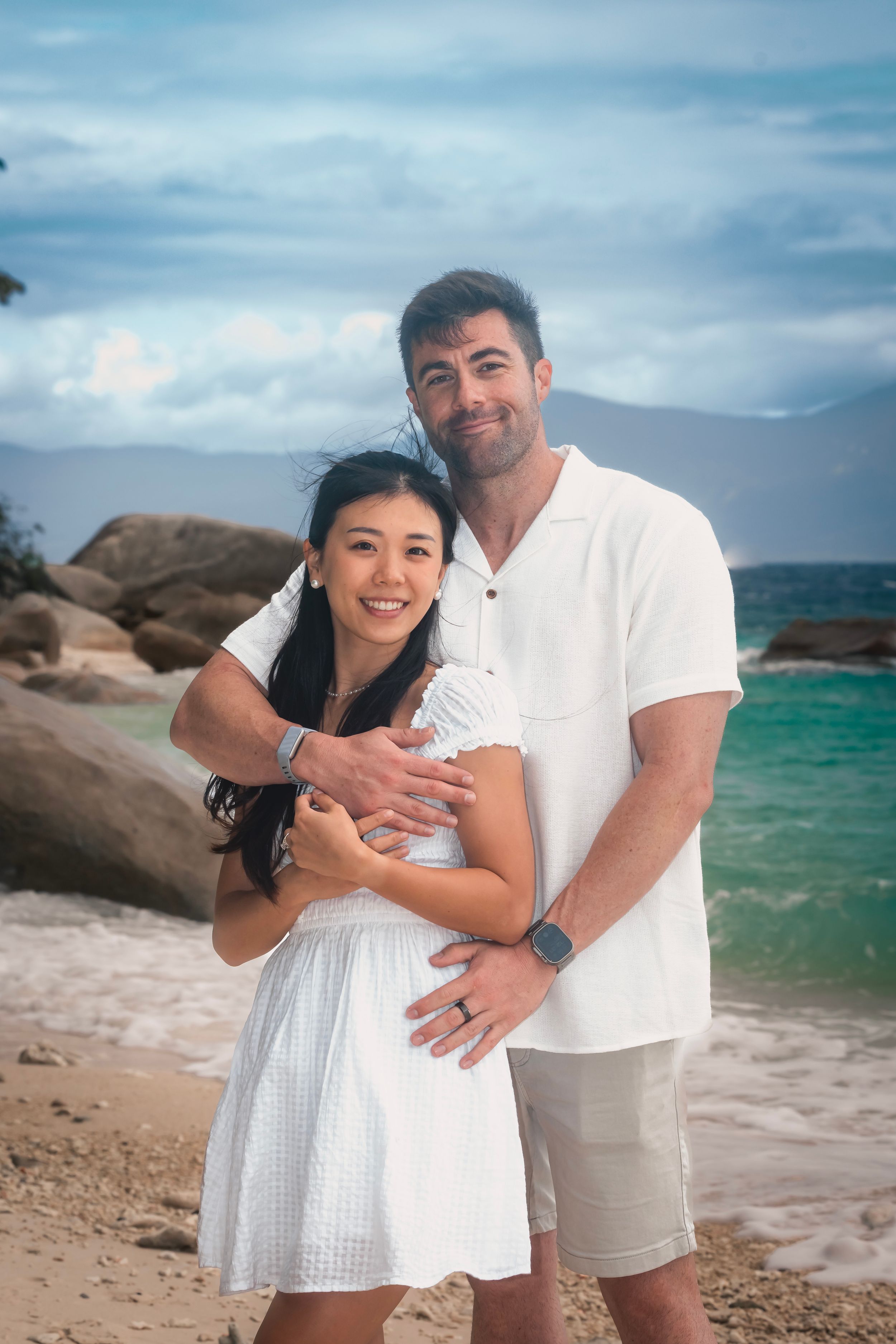 A couple embraces on a rocky beach with turquoise waters and dramatic clouds in the background.
