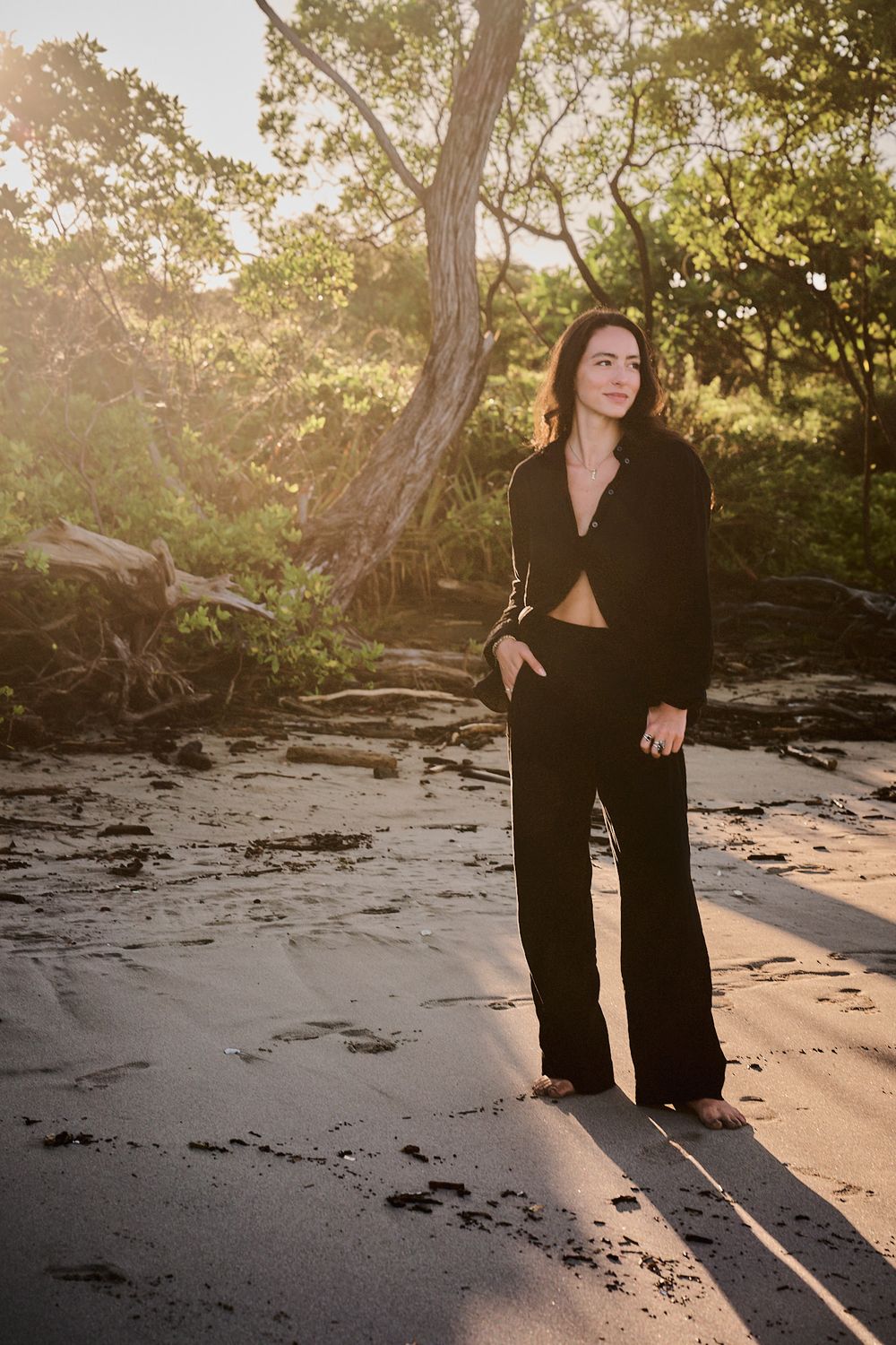 Figure in black clothing sits along rocky shoreline at sunset creating long dramatic shadows on beach.