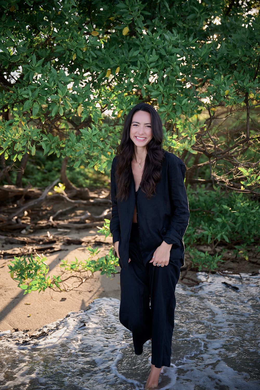A person wearing a black suit stands in front of lush greenery and a rocky stream in an outdoor setting.
