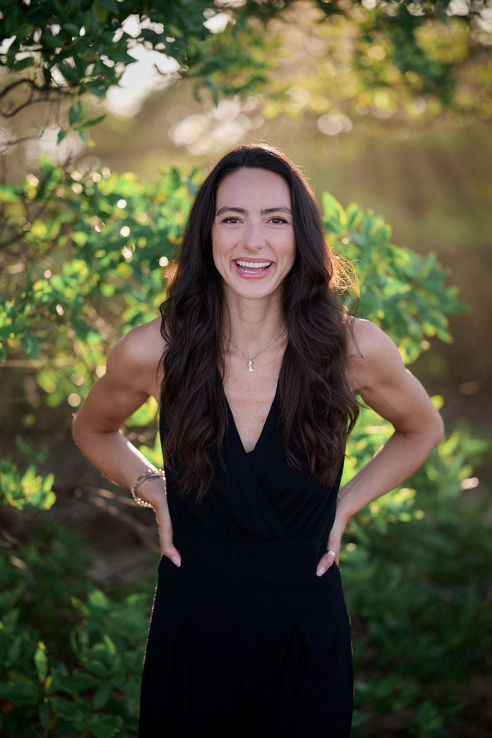 A woman in a black v-neck dress poses outdoors with hands on hips against a blurred green garden background.
