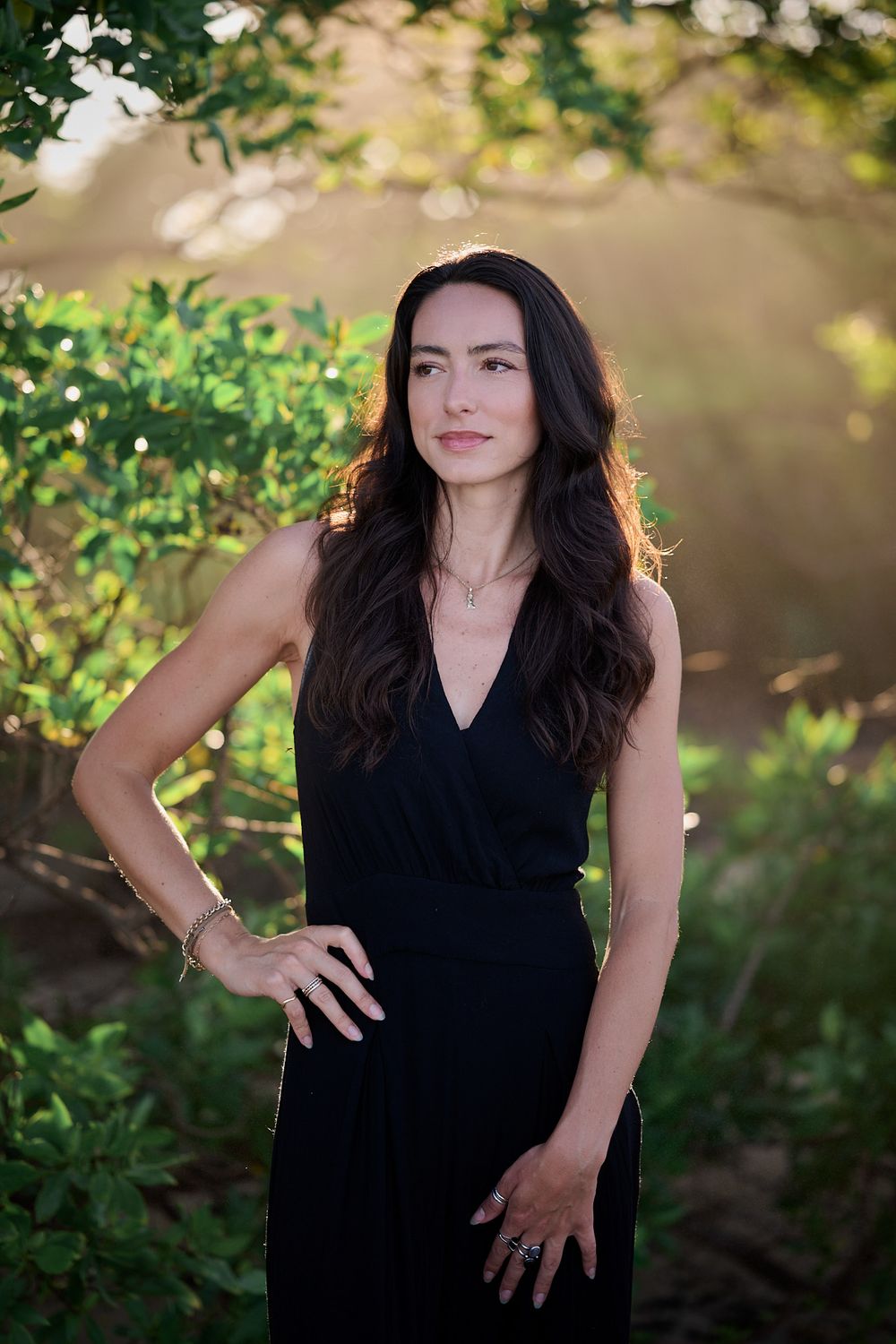 A person in an elegant black sleeveless dress poses outdoors against lush greenery and soft natural lighting.