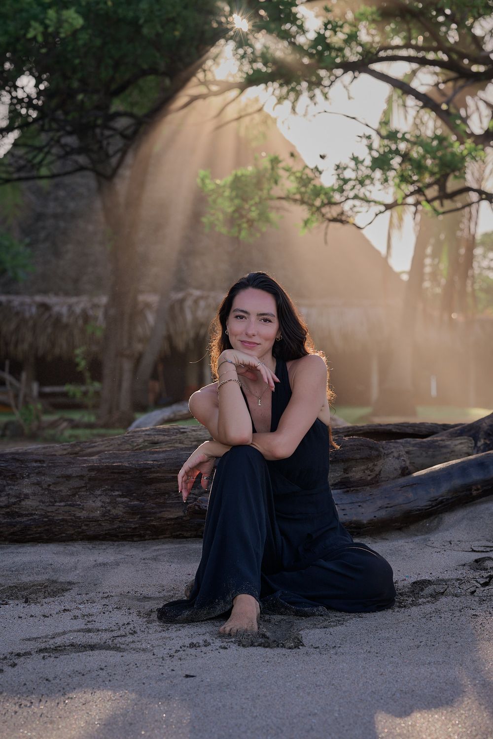 A figure in a navy dress poses gracefully in dappled sunlight beneath overhanging tree branches.