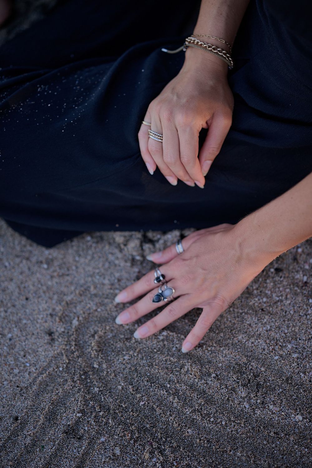 A close-up shot of hands resting on sand wearing multiple rings, showing detailed jewelry styling.