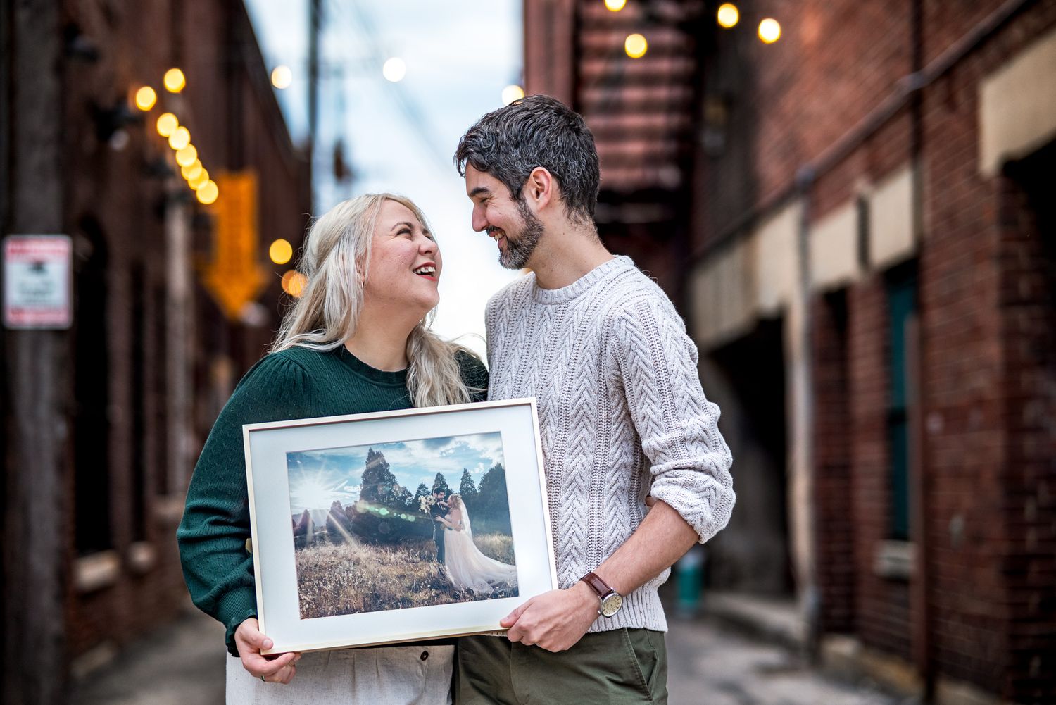A couple shares laughter while holding a framed photograph on a city street with bokeh lights in the background.