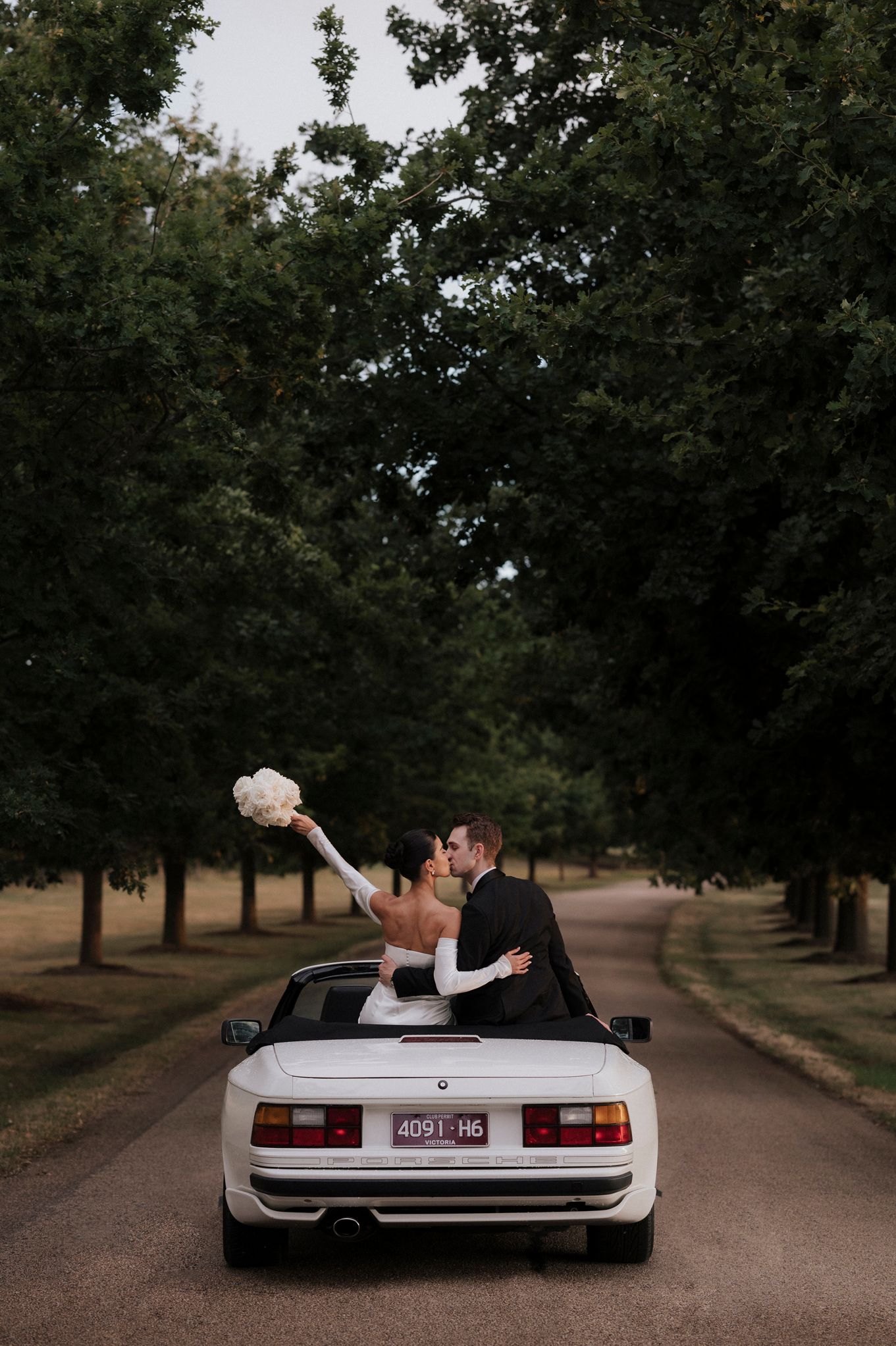 Newlyweds celebrate in white convertible car on tree-lined road.
