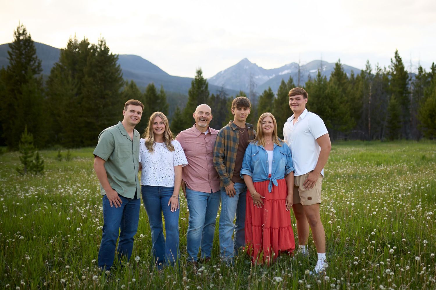 A group of people stand together in a scenic mountain meadow with wildflowers and evergreen trees in the background.