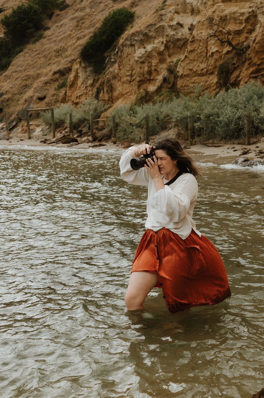 A photographer in red skirt wades into ocean water while taking pictures near coastal cliffs.