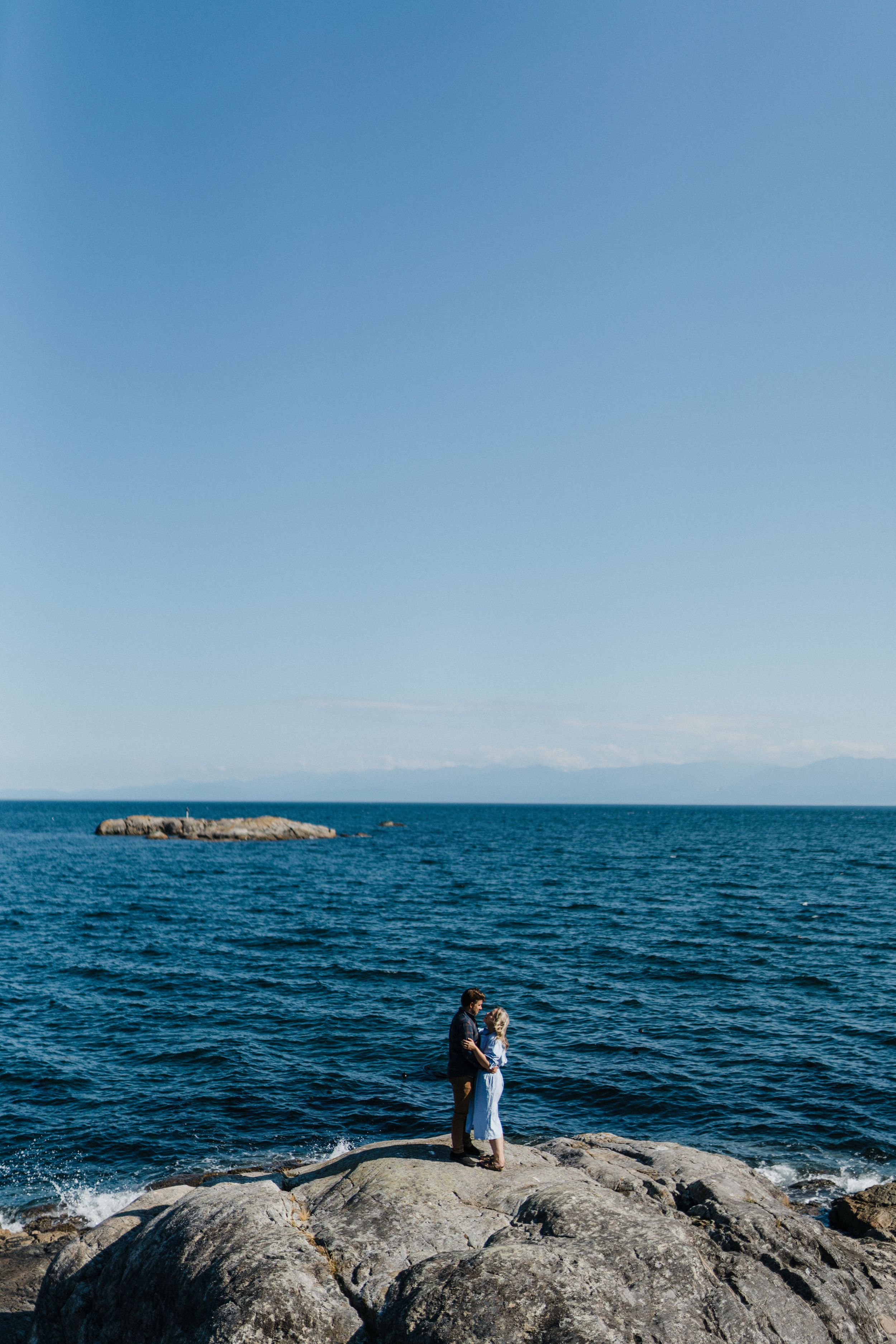 A couple stands together on a rocky outcrop overlooking the vast blue ocean on a clear sunny day.