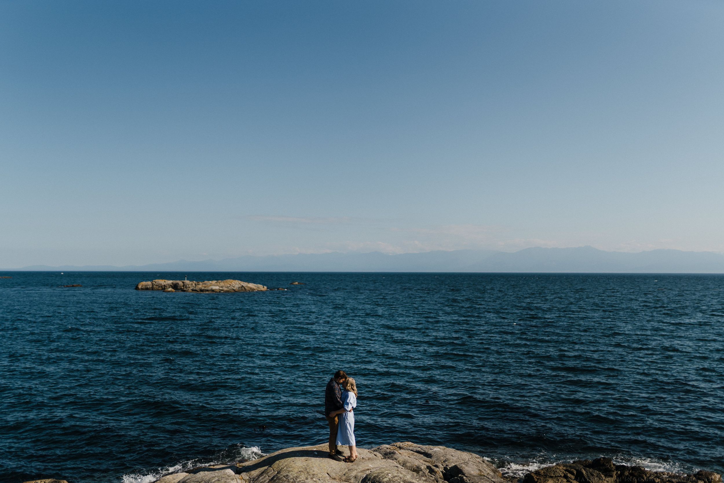 Elegant couple shares an intimate moment on a scenic coastal cliff overlooking deep blue ocean waters at sunset.