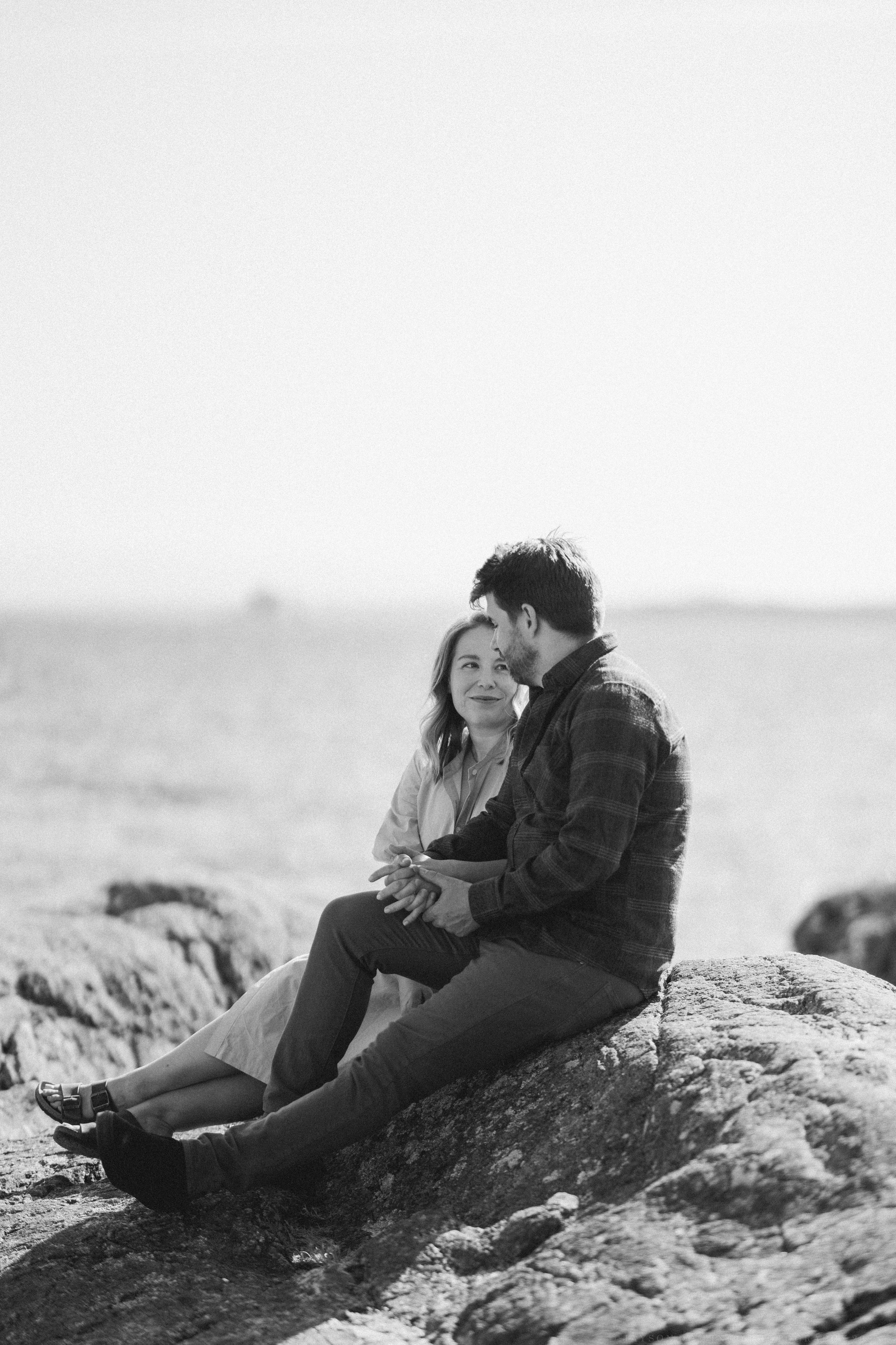 Two people relaxing together on rocky coastal cliffs in black and white.