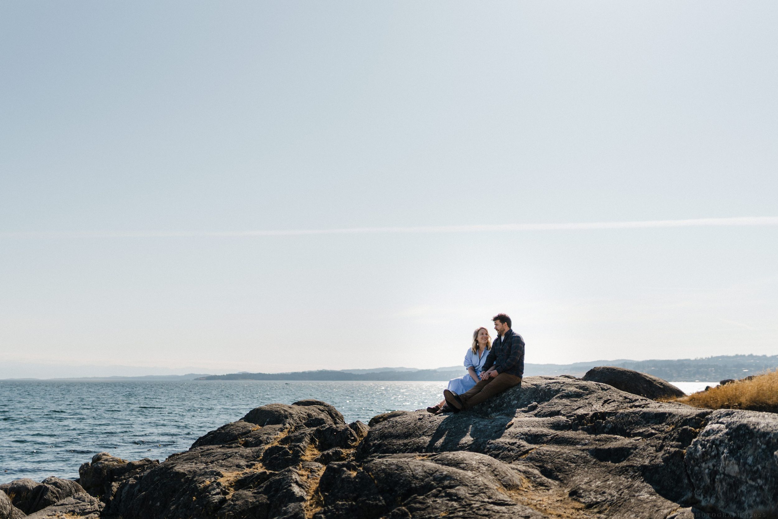 Two people sit on rocky coastal cliffs overlooking a calm ocean at sunset.