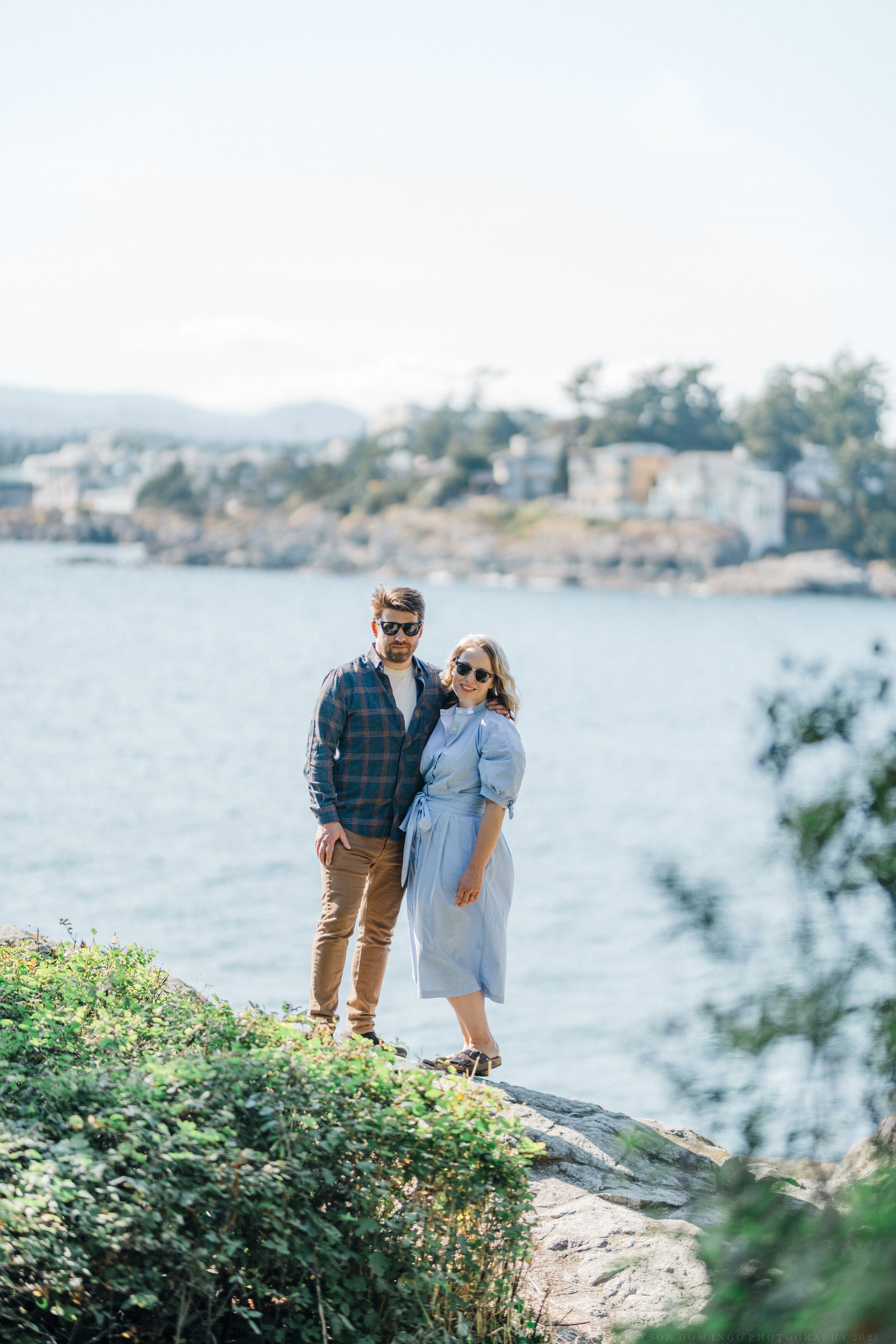 Couple enjoying scenic coastal views from a grassy cliff overlooking waterfront homes.
