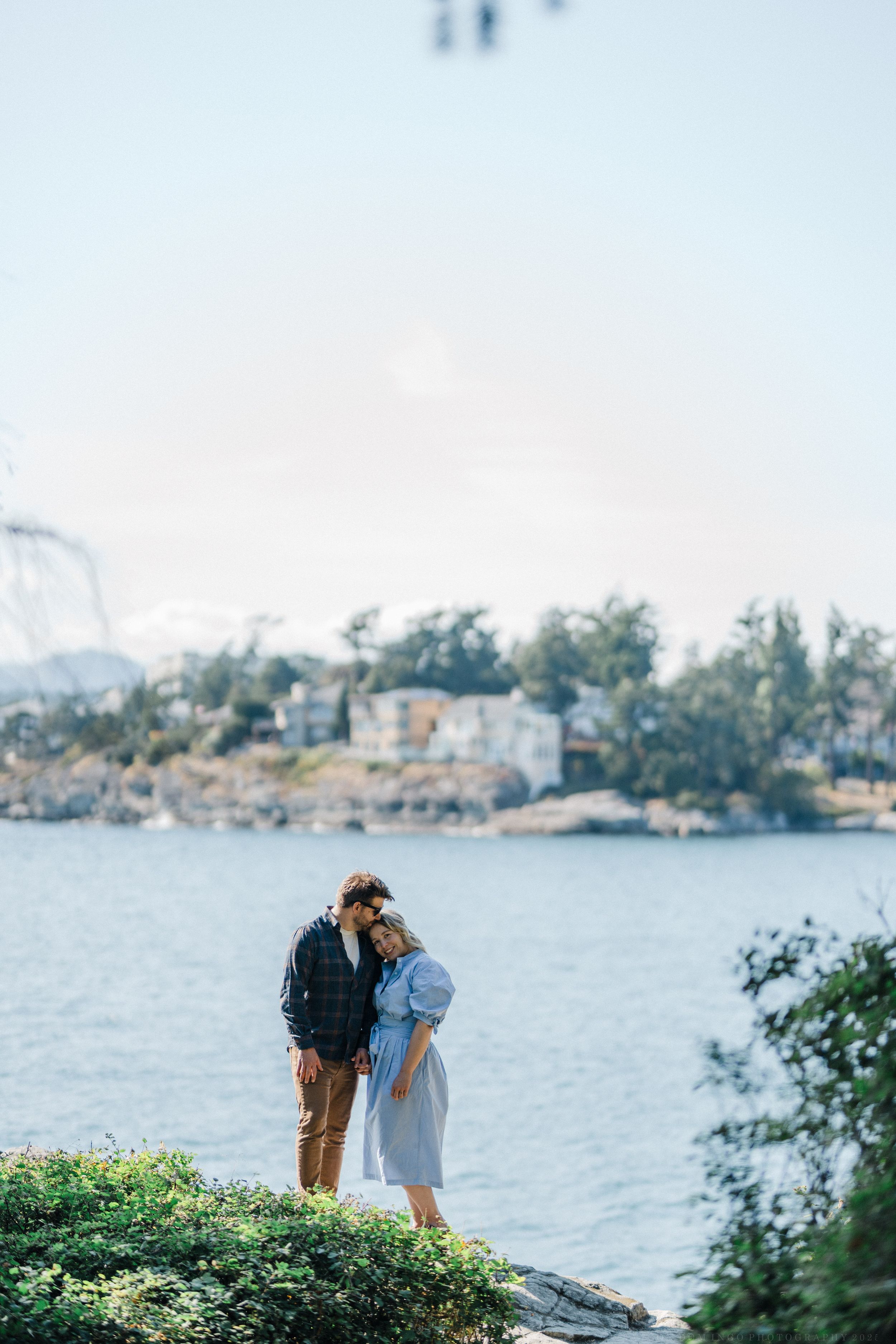 Couple sharing an intimate moment by the waterfront with boats and trees in background.