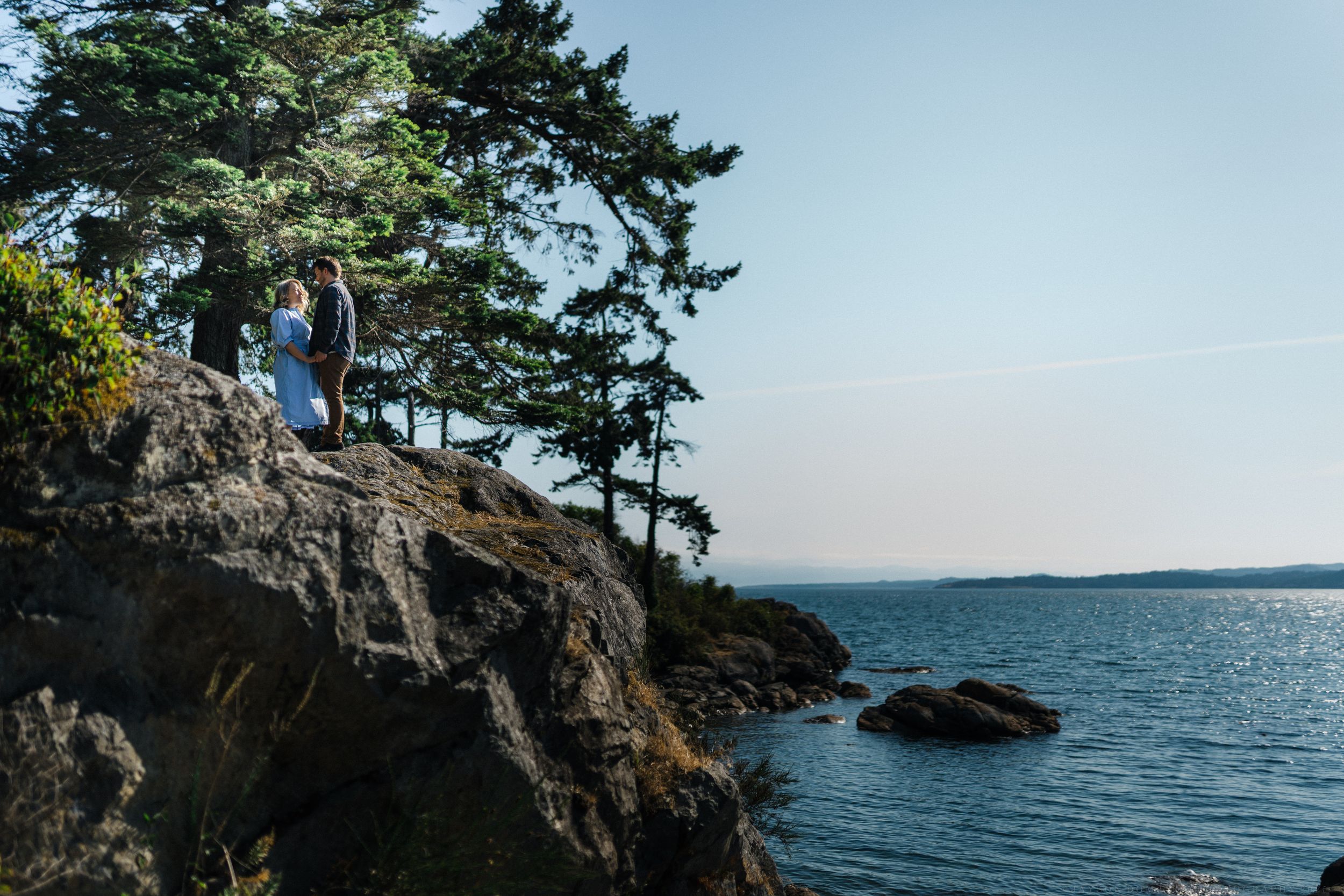 Person stands on rugged coastal cliff overlooking calm ocean waters with evergreen trees in Pacific Northwest scenery.