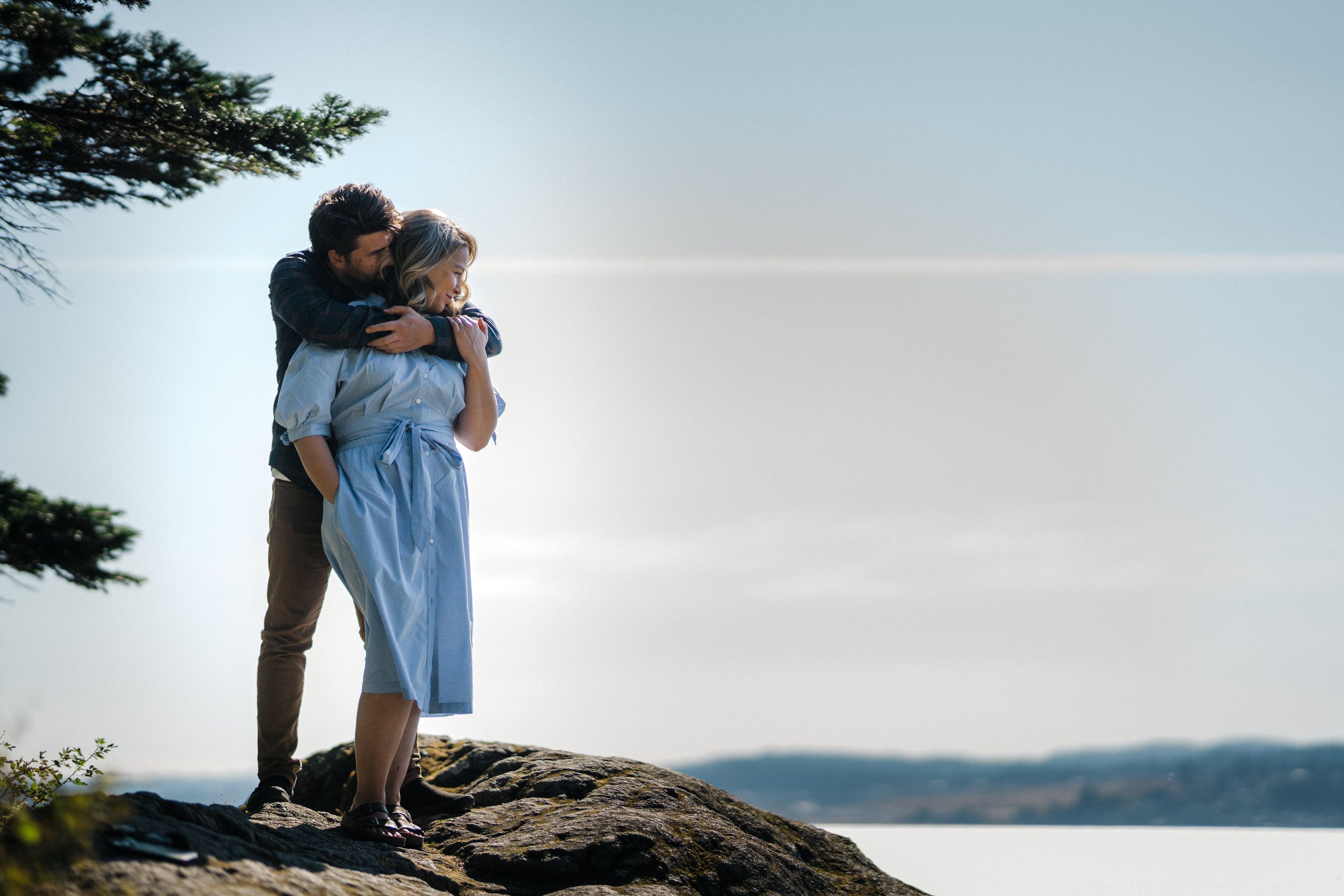 Couple embracing romantically on a rocky cliff overlooking a serene lake surrounded by trees.