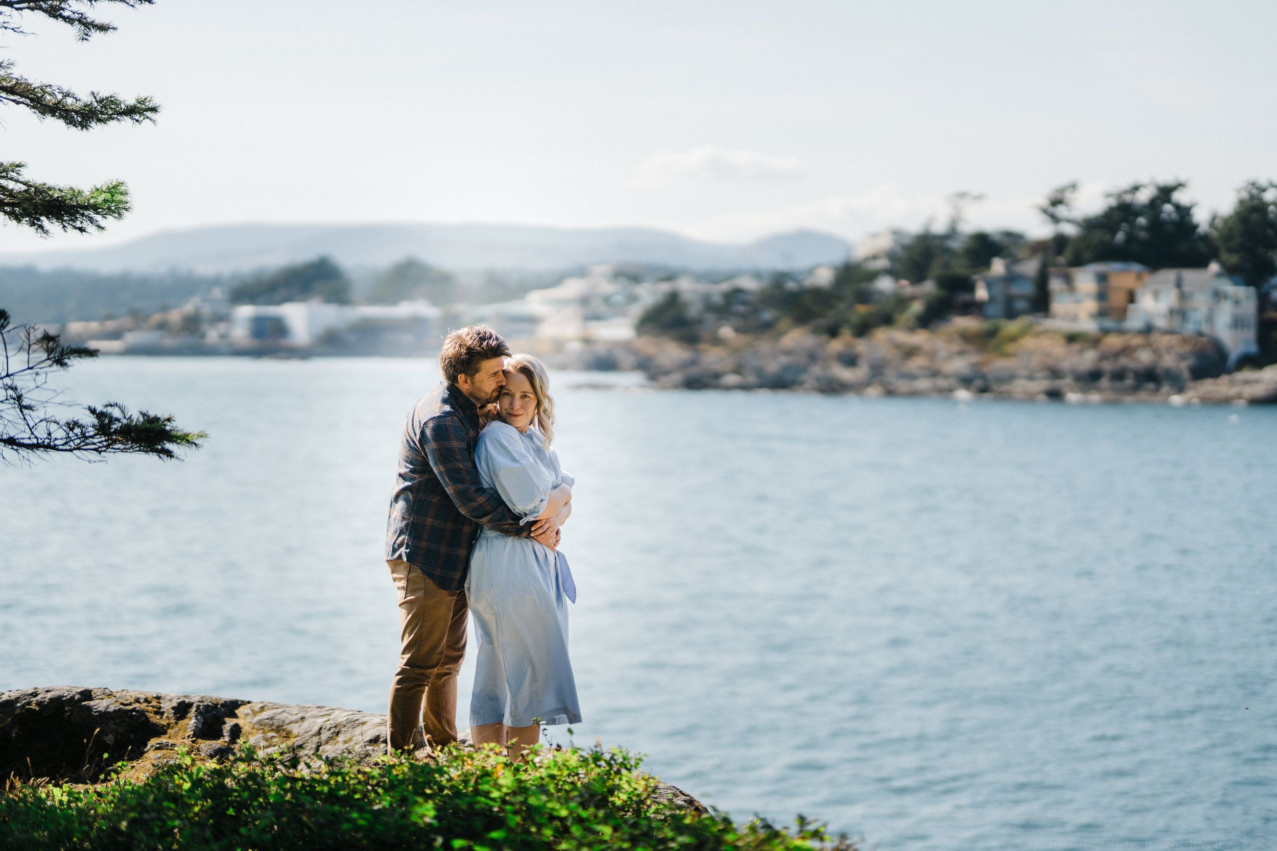 A romantic embrace on a coastal cliff overlooking a scenic bay with houses dotting the shoreline.