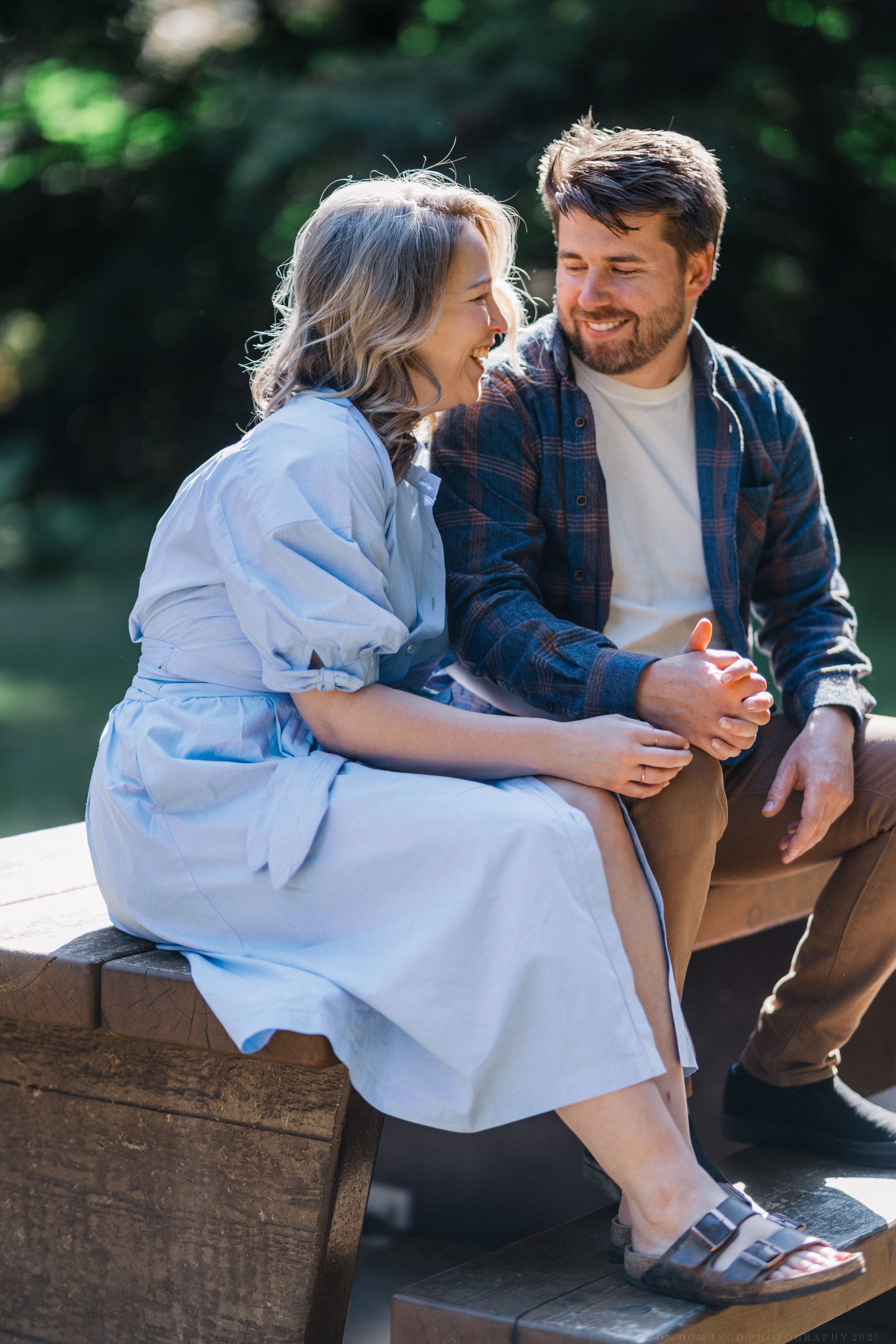 A couple shares an intimate moment while sitting on a wooden bench in a sunny garden setting.