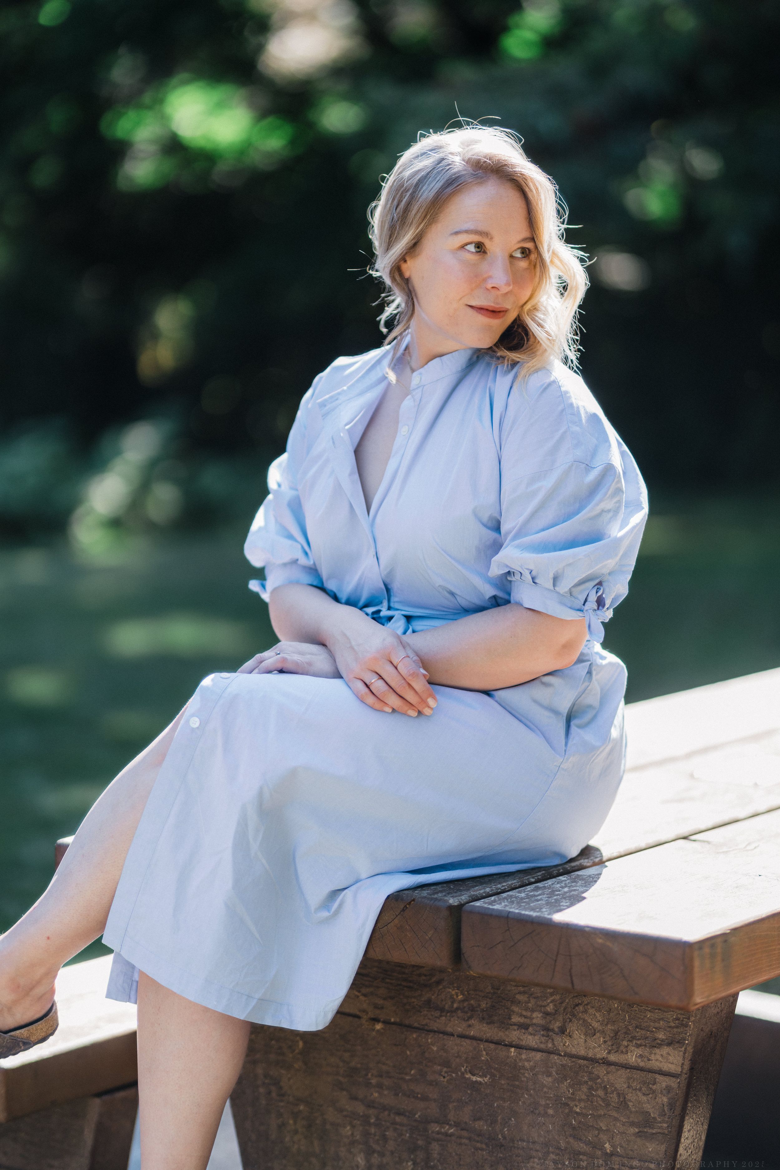 Portrait of person in light blue dress sitting on wooden bench in sunny garden setting.